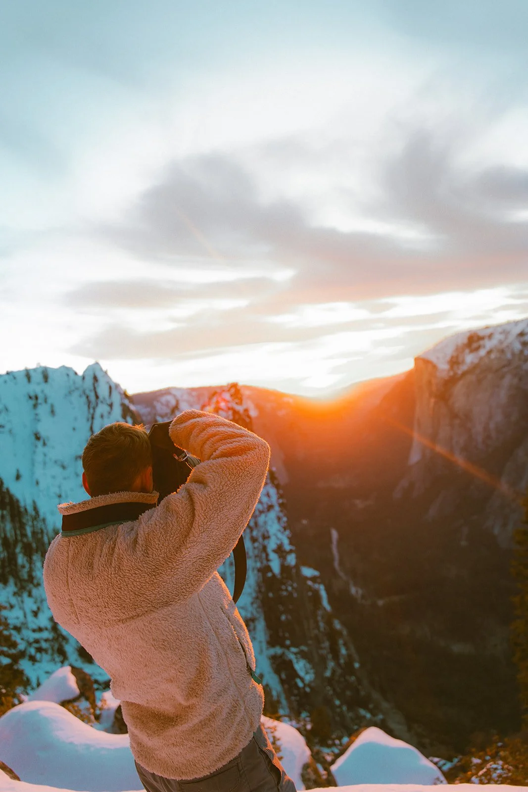 A person stands outdoors in a snowy mountainous landscape during sunset, taking a photo with a camera.