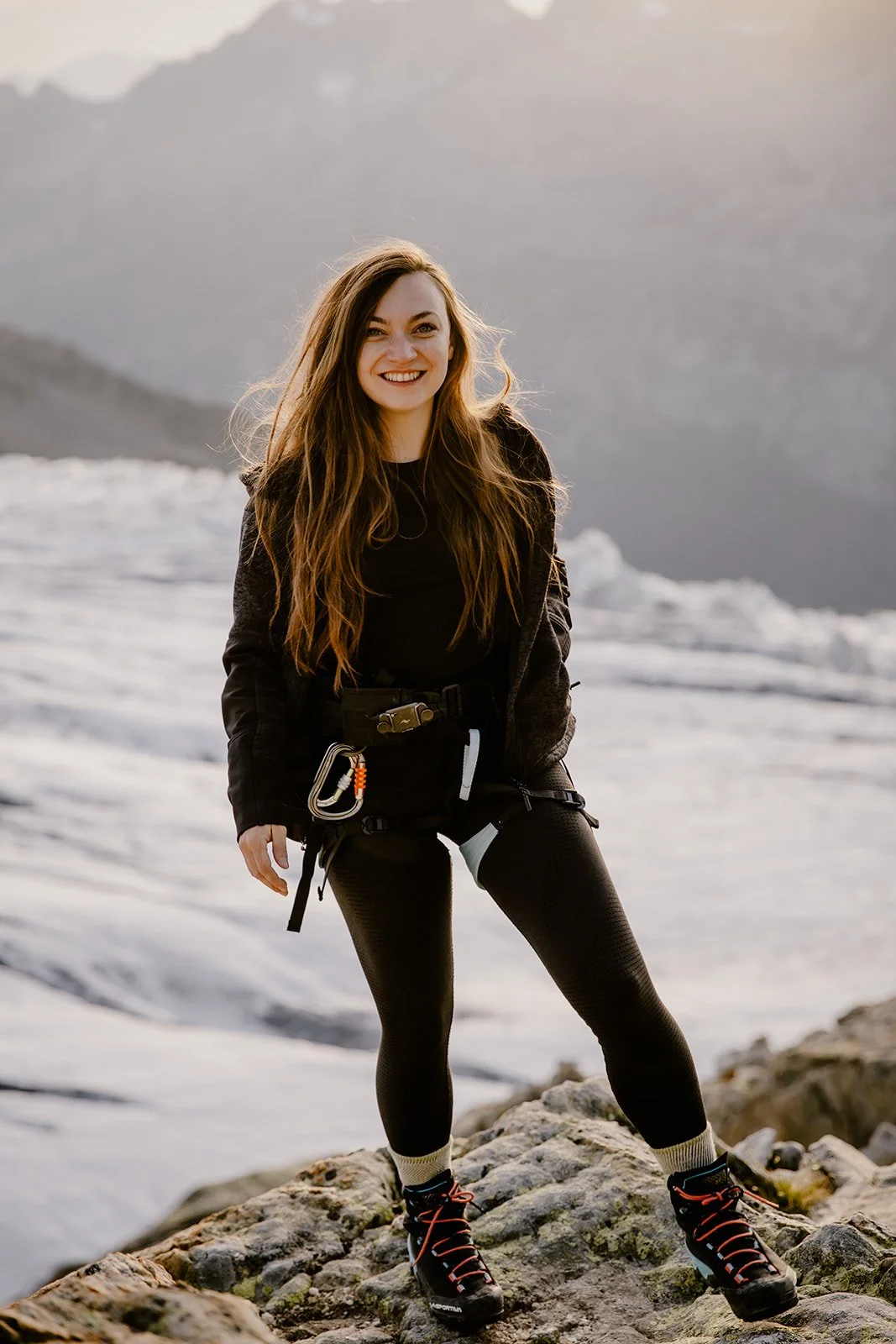 Woman standing on rocky terrain near water, smiling, wearing outdoor clothing and hiking boots.