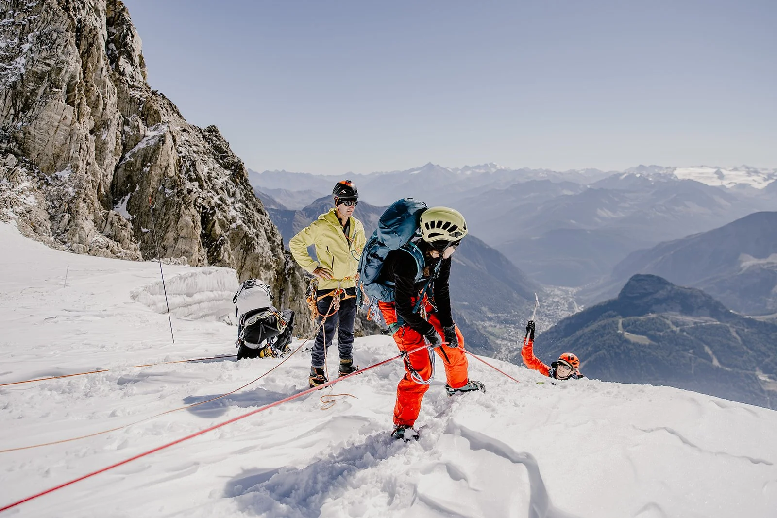 Mountaineers climbing a snowy slope in the mountains, with one fallen and others helping or resting, mountain landscape in the background.