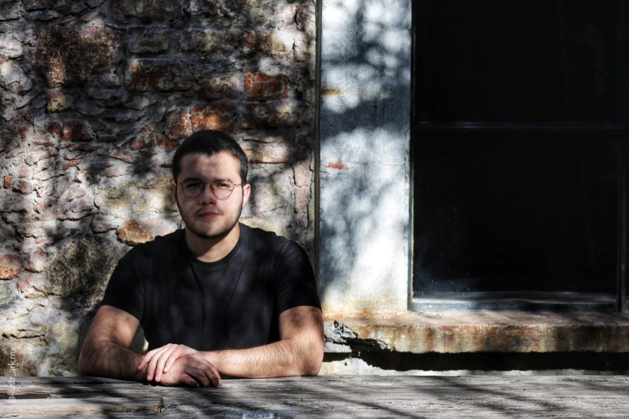 Joven con gafas sentado en una mesa de madera frente a una pared de ladrillos y un ventanal con sombra de árboles.