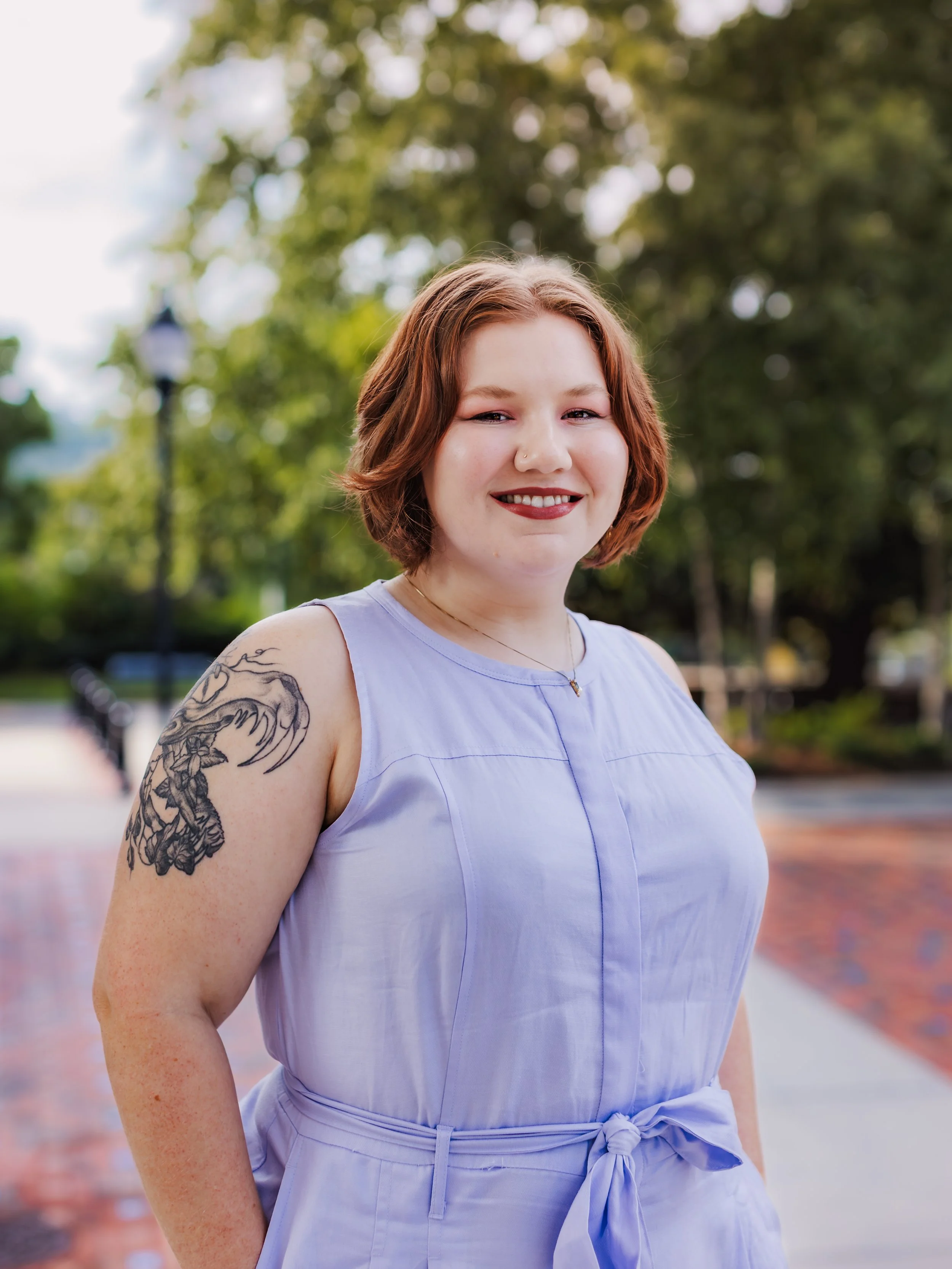 Madison Root, founder of Lavender Root Legal, with short red hair and a tattoo on her left shoulder, wearing a light purple sleeveless dress, smiling outdoors in downtown Asheville with trees and a brick-paved walkway in the background.