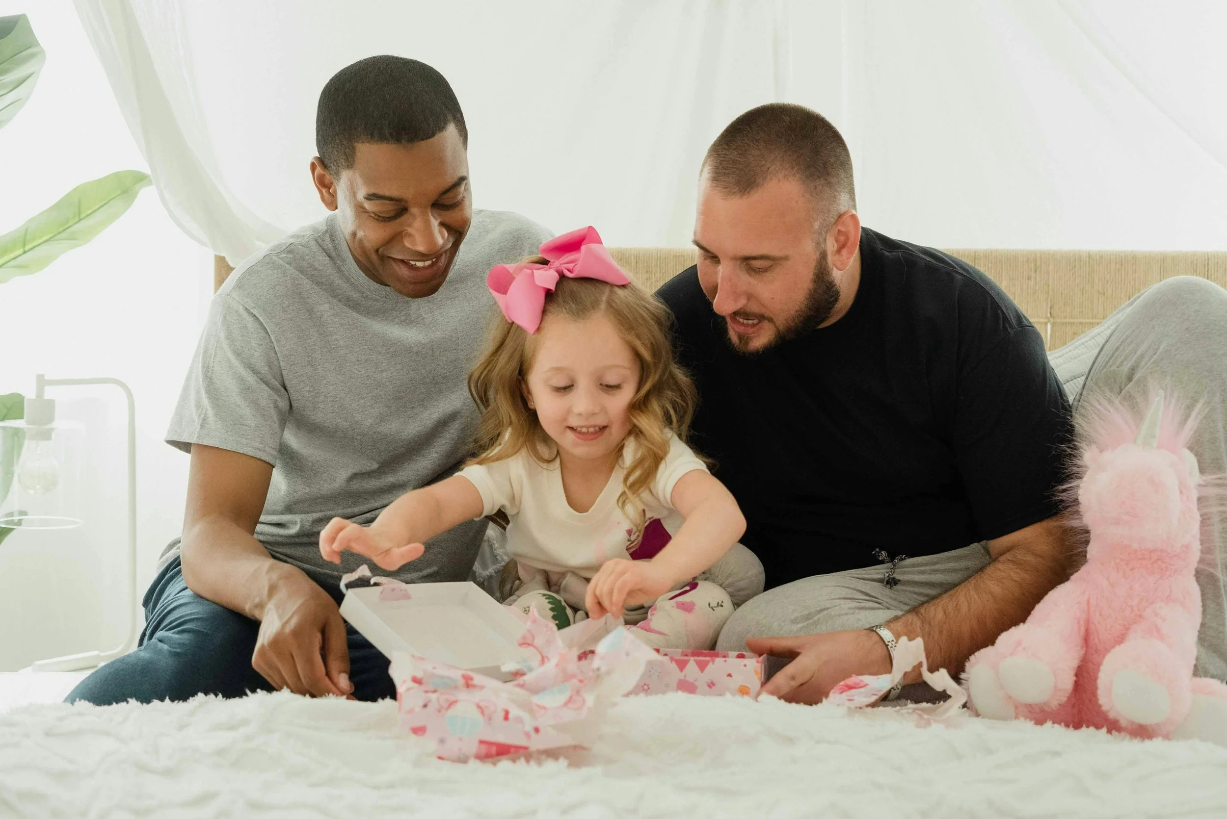 A young girl with a pink bow opens a gift box between by two men all sitting on the girl's bed with her plush pink unicorn toy, in a bright room.