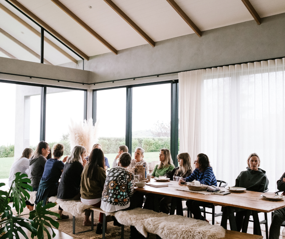 A group of people sitting around a long table in a well-lit room with large windows and white curtains, engaging in a discussion or meeting.