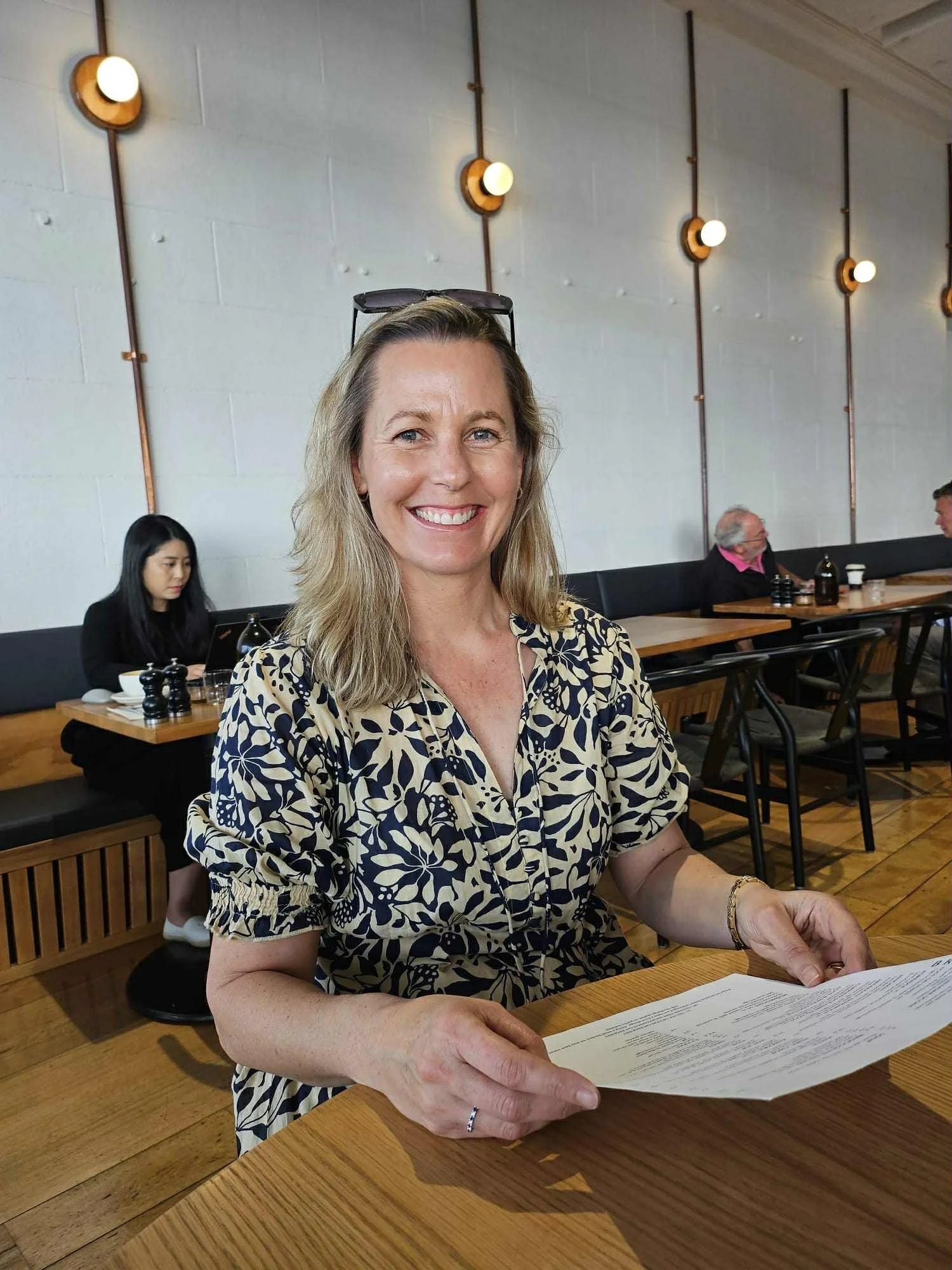 A smiling woman with blonde hair wearing a patterned dress sits at a wooden table in a modern cafe, holding a menu. The cafe has a white wall with black and copper light fixtures, and other patrons are visible in the background.