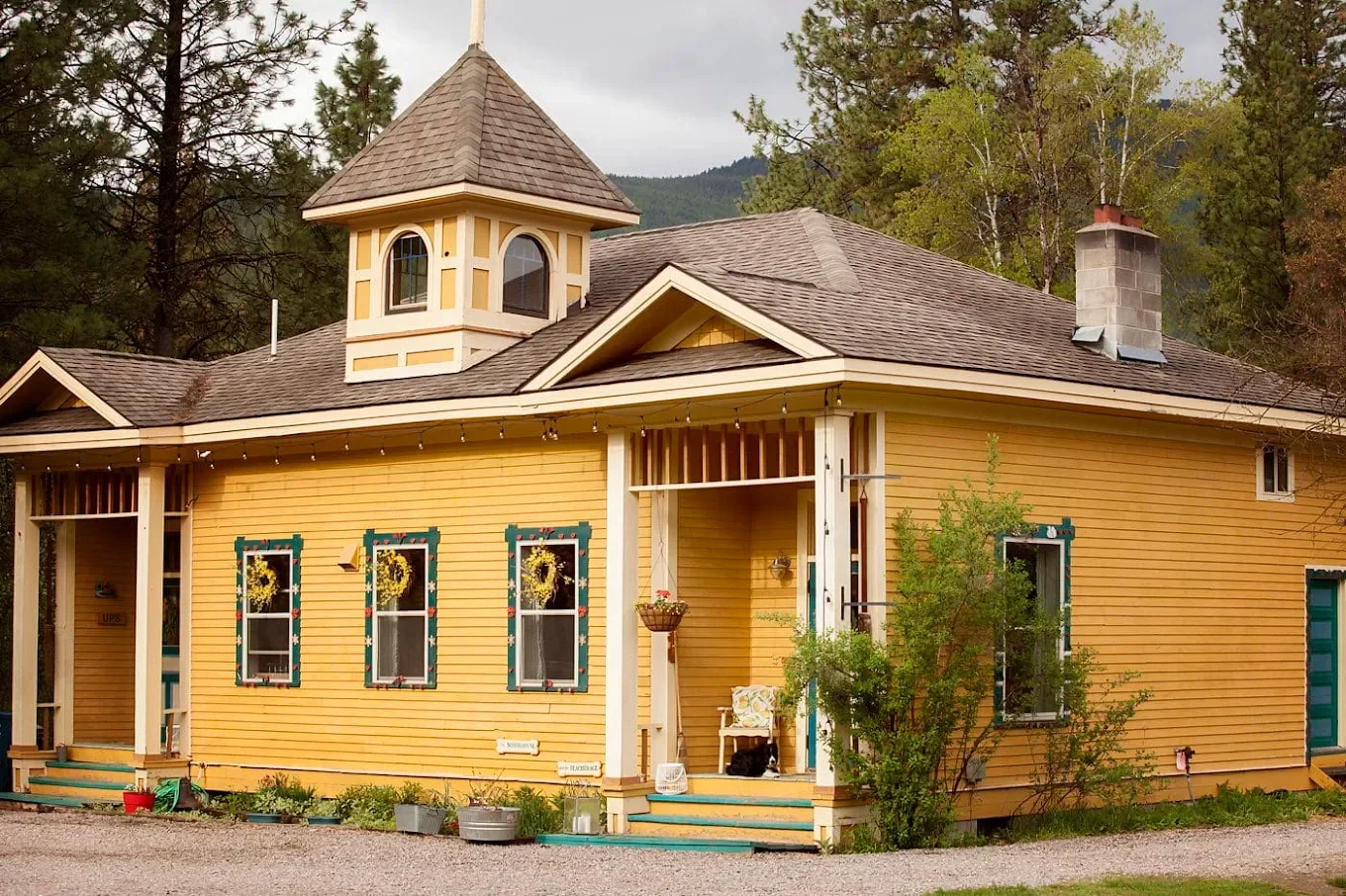 A yellow house with decorative window frames and a small front porch, surrounded by greenery and trees, with a mountain in the background.
