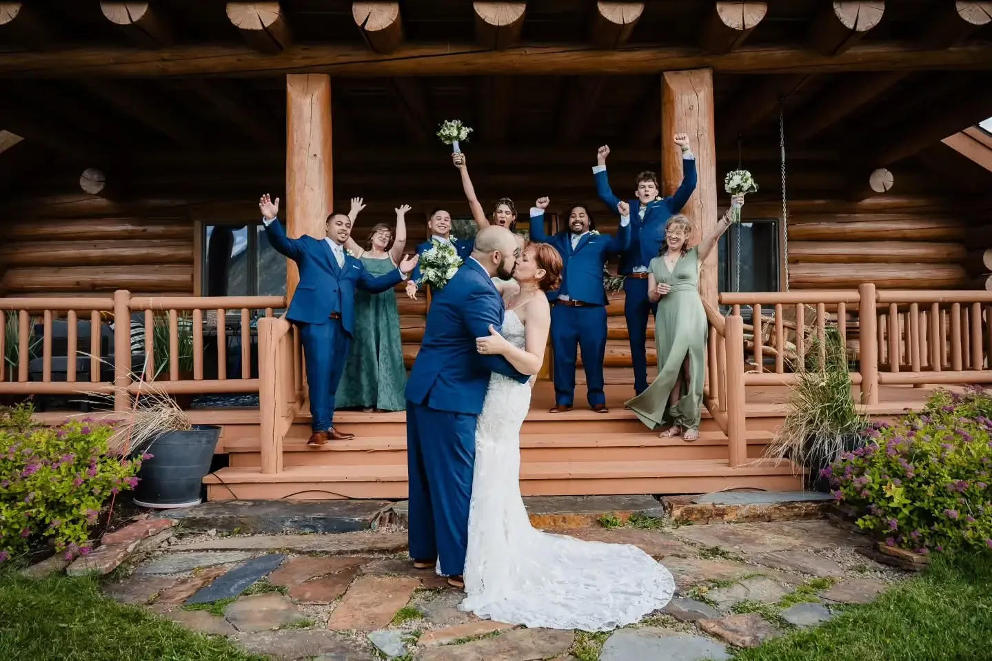 A wedding couple is kissing in front of their wedding party on a wooden porch, with the bridesmaids and groomsmen celebrating behind them, some raising their arms and holding bouquets.