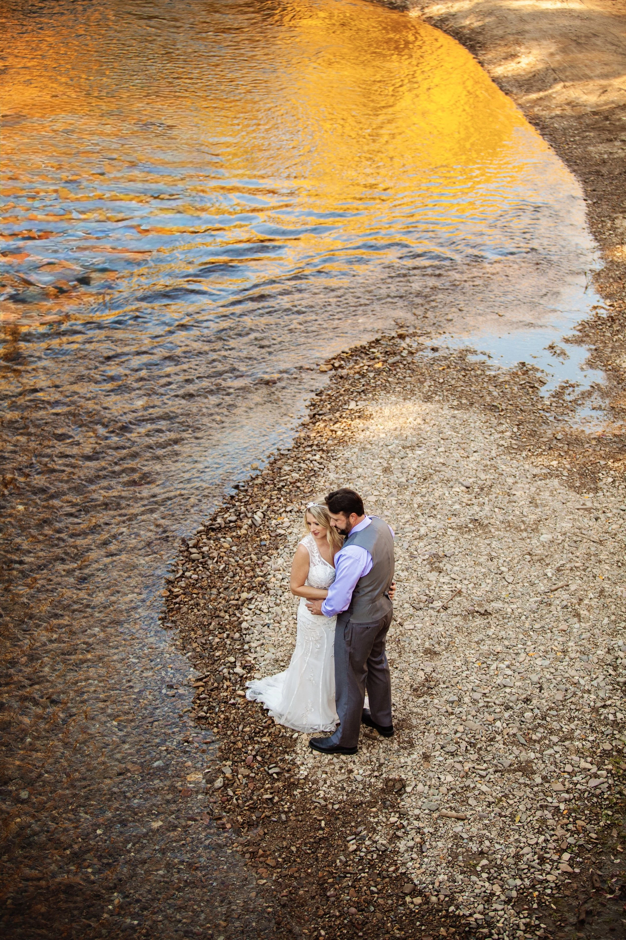 A bride and groom hugging on a rocky beach near the water at sunset.