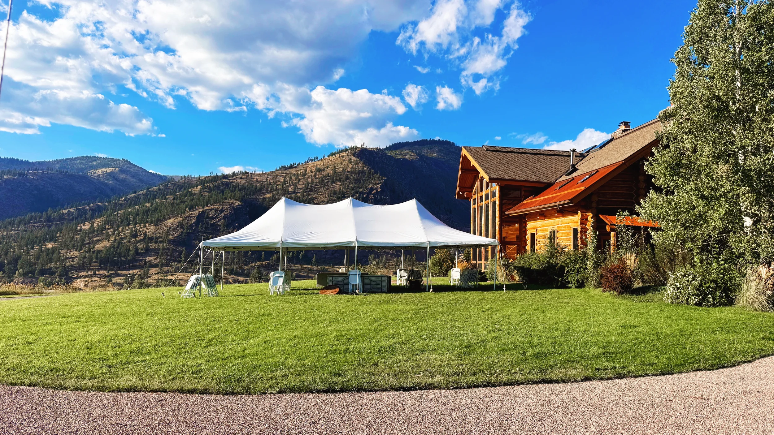 A large white event tent set up on a lush green lawn in front of a log cabin with mountains in the background, under a partly cloudy blue sky.