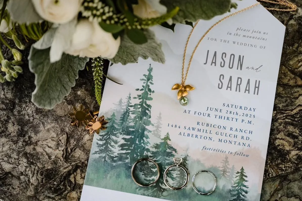 Wedding invitation with greenery, flower jewelry, and rings on a tree bark background. The invitation is for Jason and Sarah's wedding on June 28, 2025, at 4:30 PM, at Rubicon Ranch, Alberta, Montana.