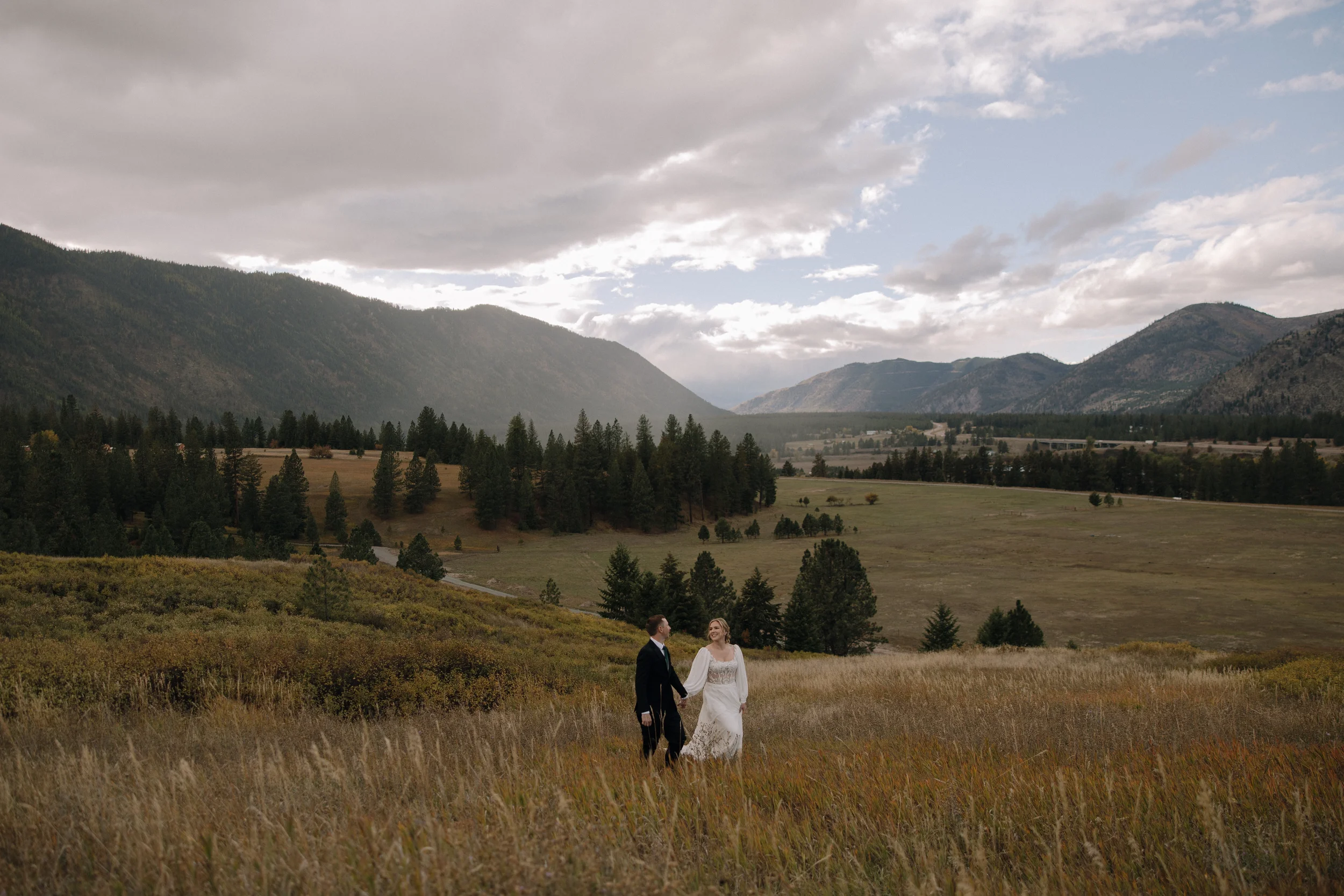 A bride and groom walking hand in hand in a grassy field with a mountainous landscape and cloudy sky in the background during daytime.