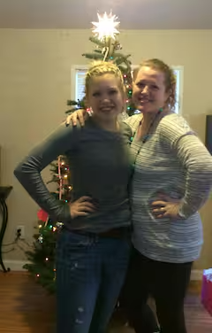 Two women standing indoors in front of a Christmas tree with a star topper, smiling with arms around each other.