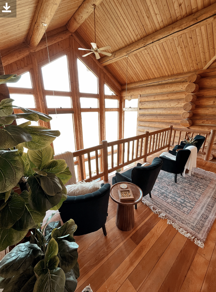 Interior of a log cabin with a wooden ceiling, large windows, and a sitting area with black armchairs, a small wooden side table, and an area rug.