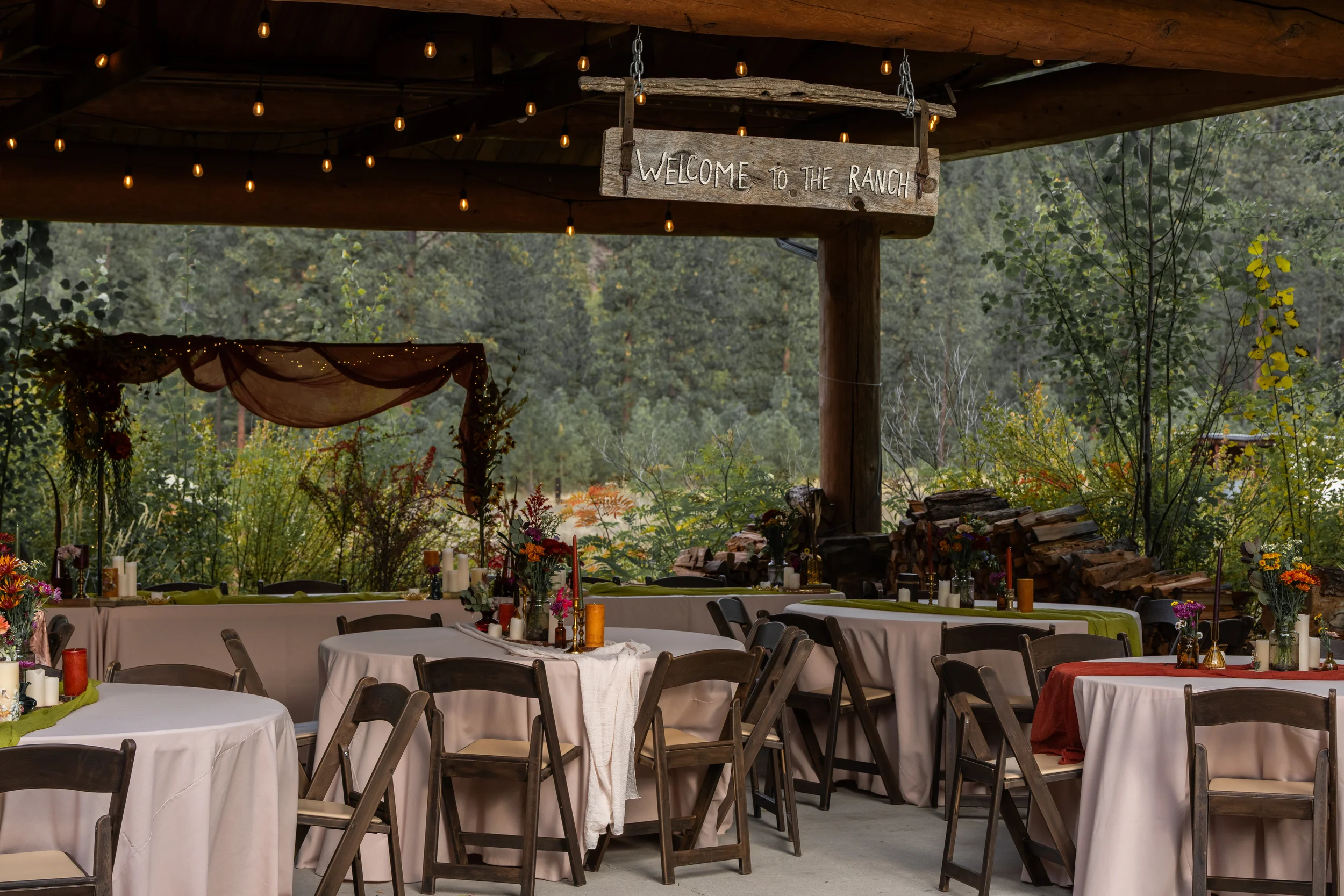 Outdoor rustic wedding reception with round tables covered in white and red tablecloths, decorated with flowers and candles, under a wooden canopy with a sign that reads "Welcome to the Ranch" and string lights, set against a backdrop of trees and stacked firewood.