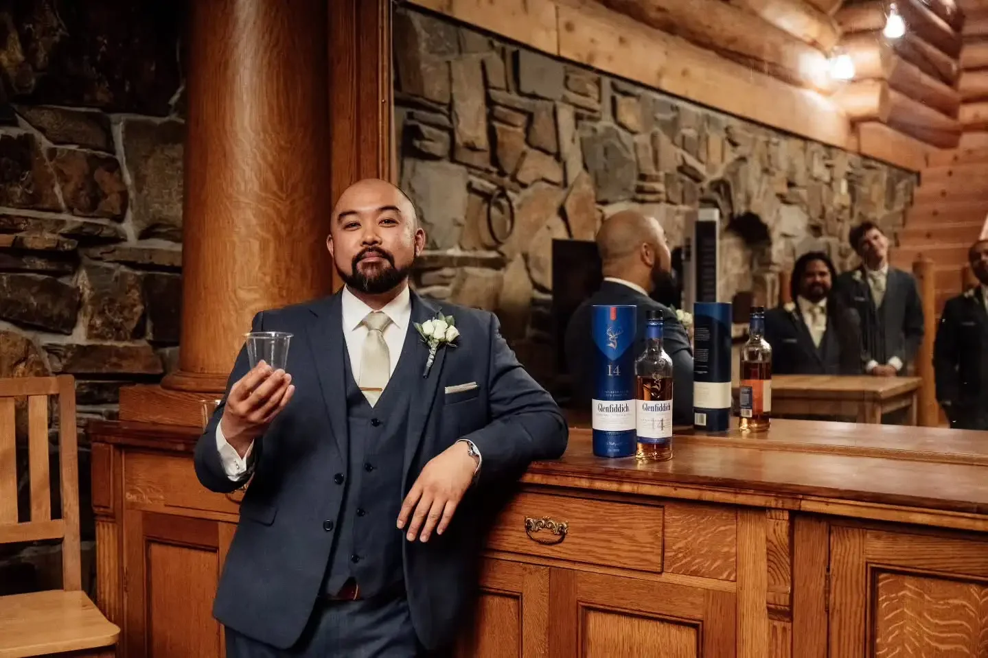 A man in a suit holding a clear glass, standing at a bar counter in a rustic wooden and stone lodge, with four men in suits in the background.