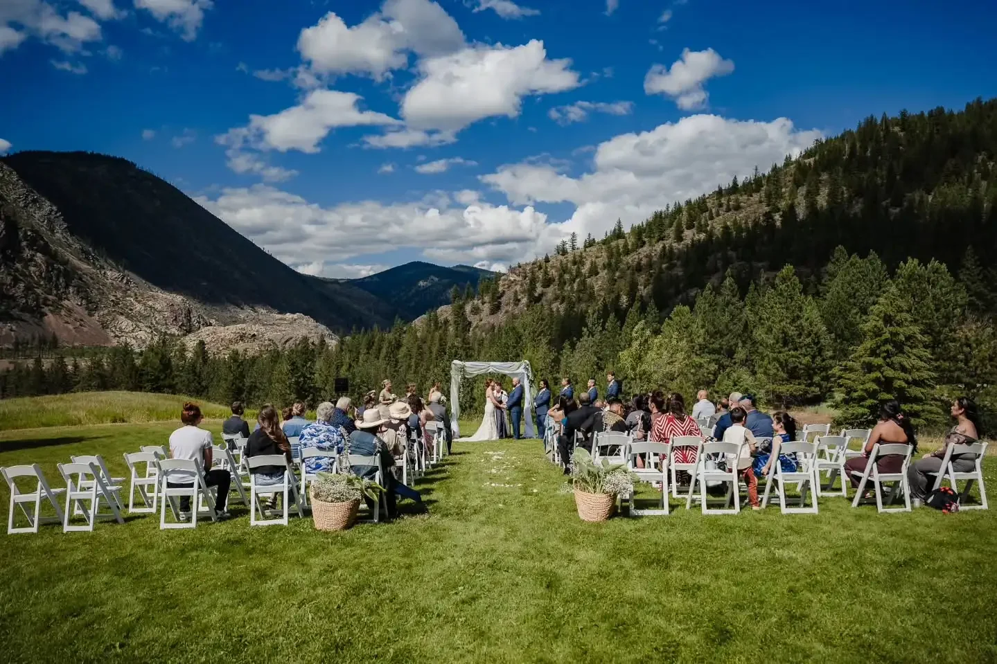Outdoor wedding ceremony set in a lush green field surrounded by mountains and pine trees, with a white arch and guests seated on white chairs.