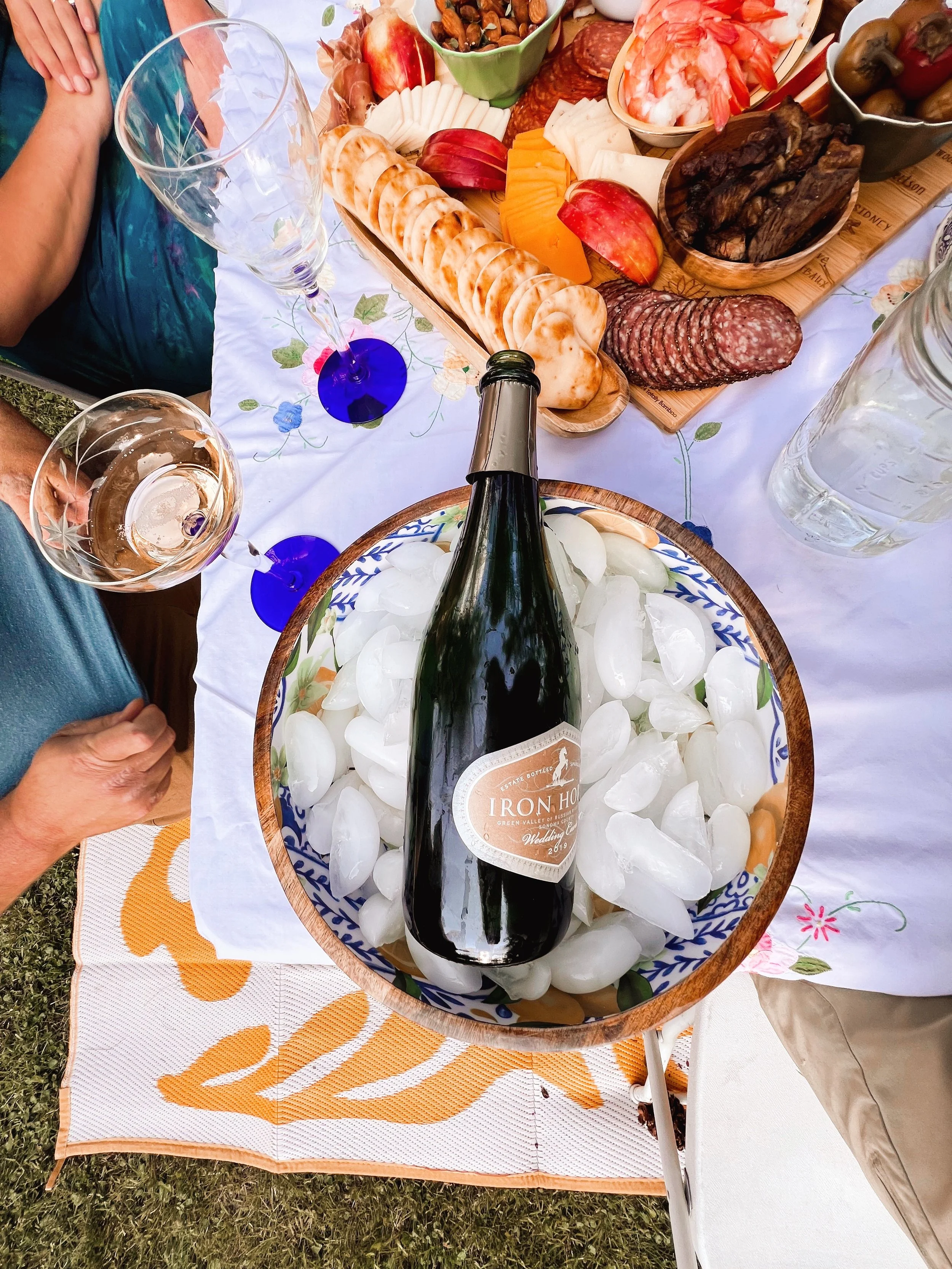 A picnic table with a charcuterie platter, cheese, bread, fruit, and a bottle of wine chilling on ice, surrounded by glasses and people.