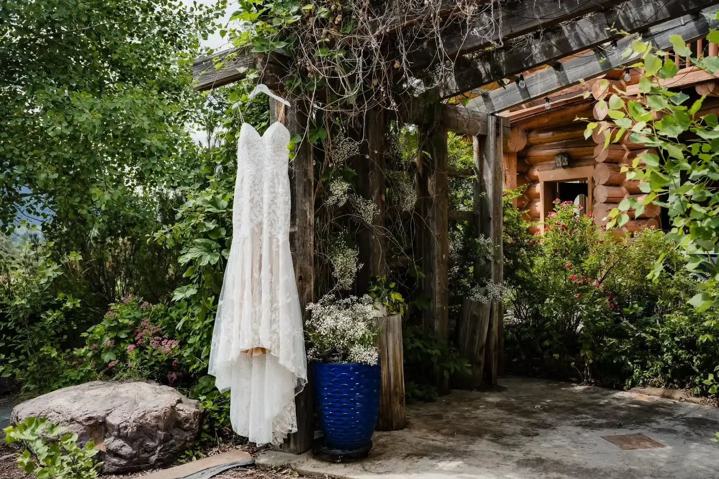 White wedding dress hanging on a hanger, placed outside among greenery, next to a blue vase with white flowers, with a wooden log cabin visible in the background.