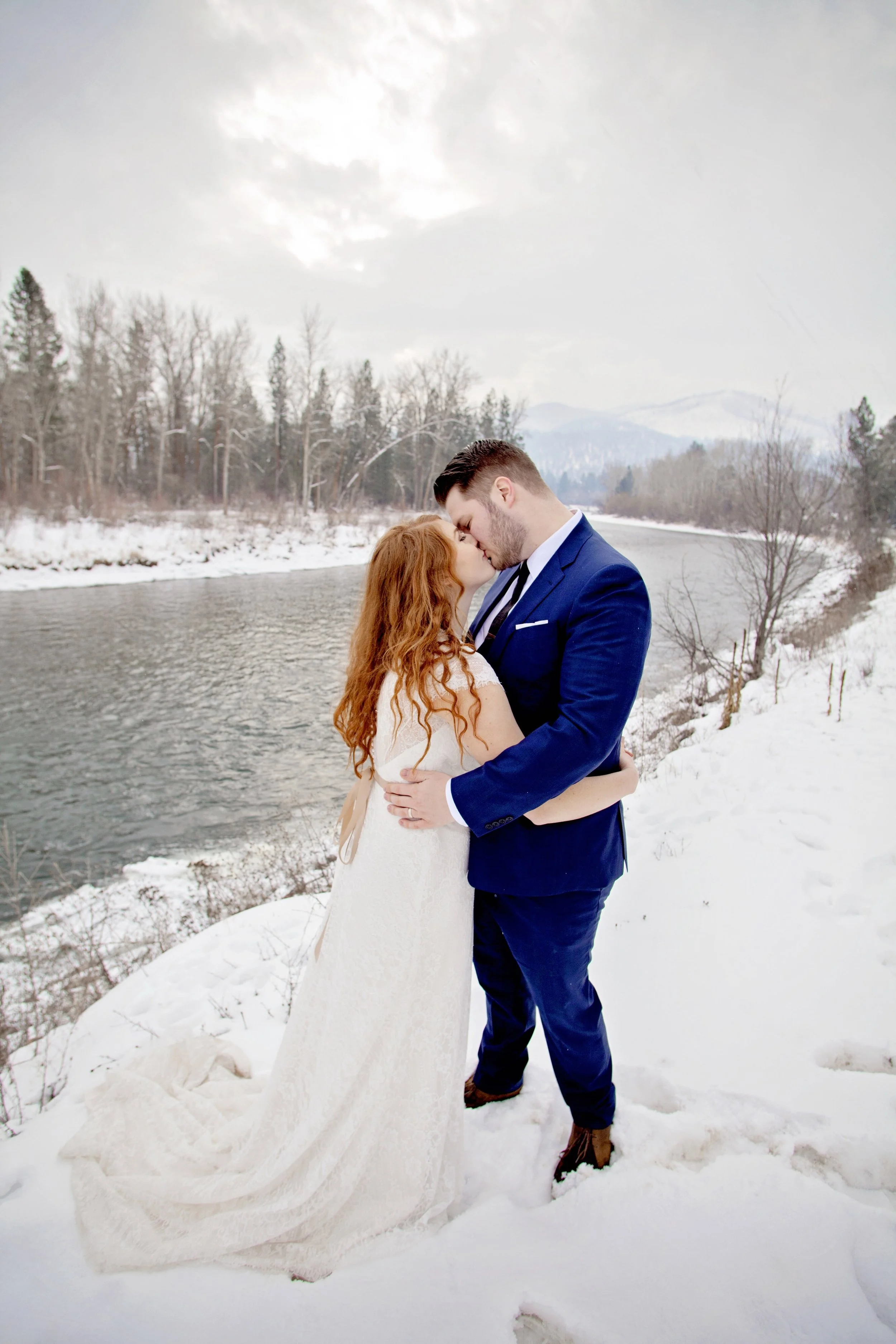 A couple in formal attire sharing a kiss in a snowy outdoor setting near a river, with snow-covered trees and mountains in the background.