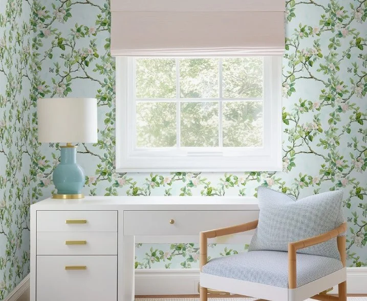A window with a light pink shade, floral wallpaper with green leaves and pink flowers, a white desk with gold handles, a blue table lamp, and a light blue armchair with a gray pillow.