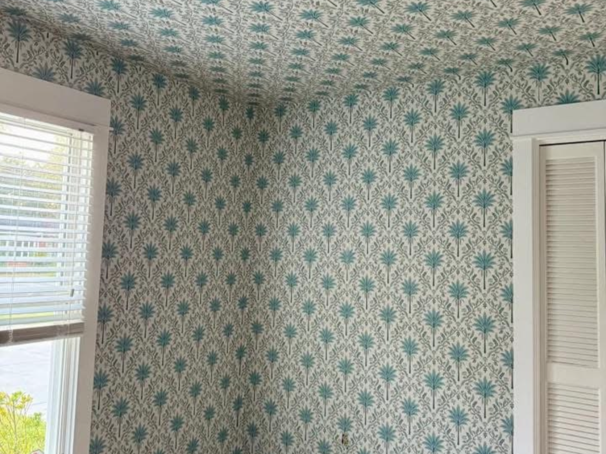 A hotel room with a large bed, decorative pillows, a sofa in front, a desk with a chair, a lamp, and patterned wallpaper with a branch and leaf design.