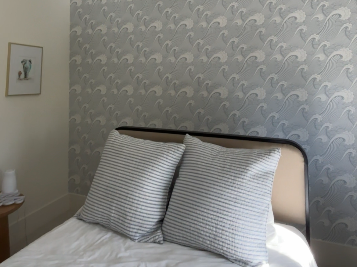 Decorative indoor space with a green leafy patterned wallpaper, floating shelves holding potted plants, books, and pottery, and a radiator with magazines and cords on top.