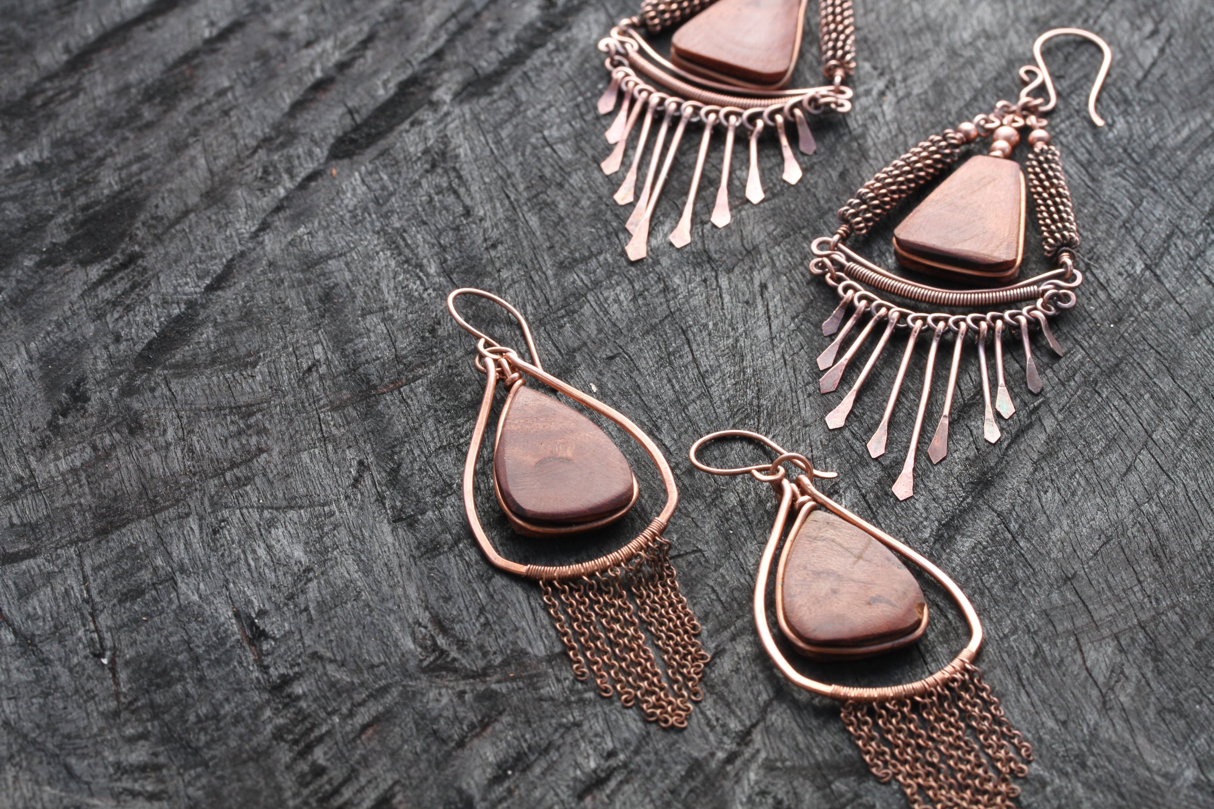 Pair of statement earrings with triangular dark timber stones, copper wire work, and dangling chains, arranged on a textured dark grey surface.