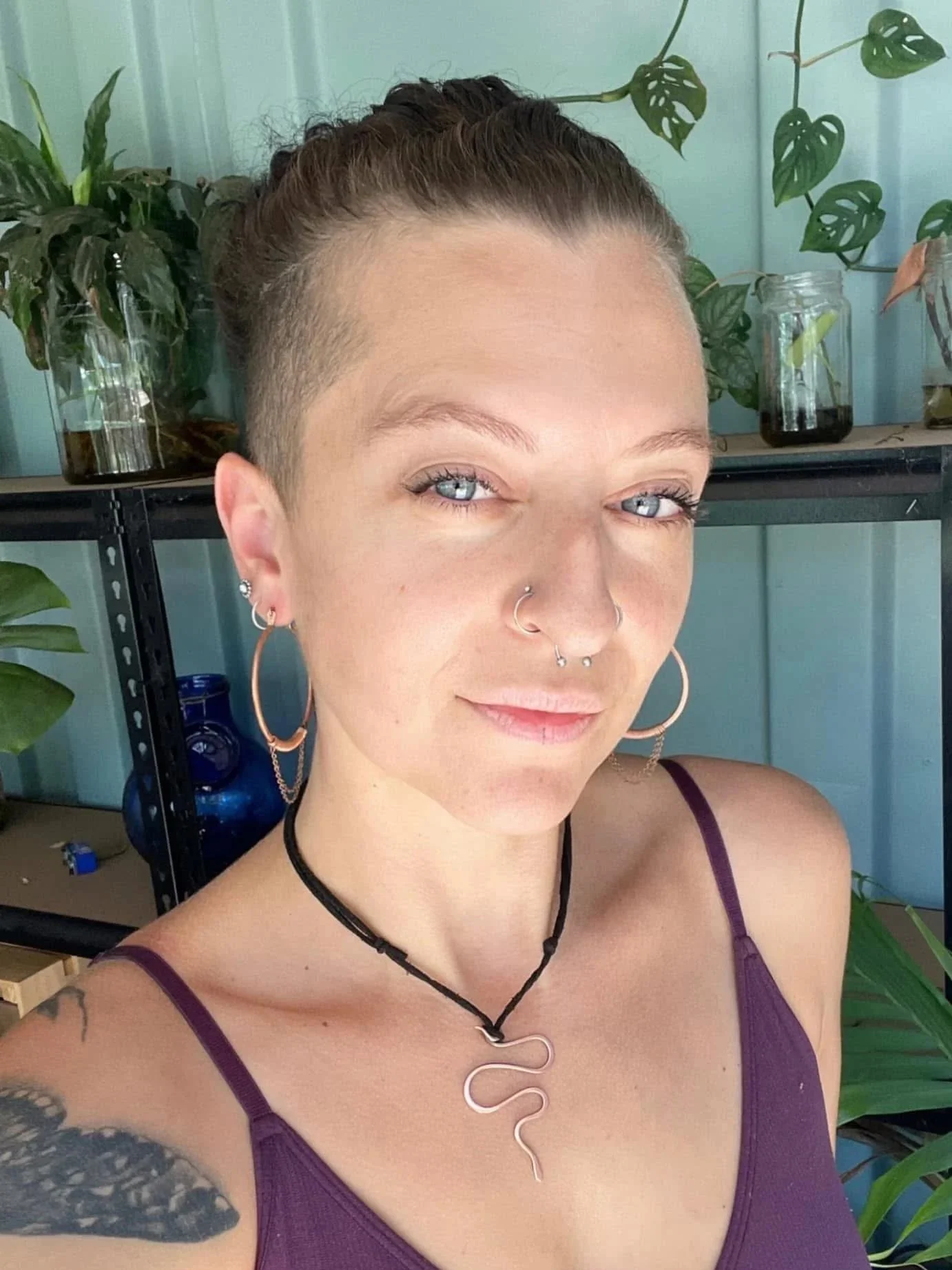 A woman with short hair, multiple facial piercings, blue eyes, and tattoos, wearing a purple tank top and a snake-shaped necklace, takes a selfie indoors with plants and glass jars in the background.