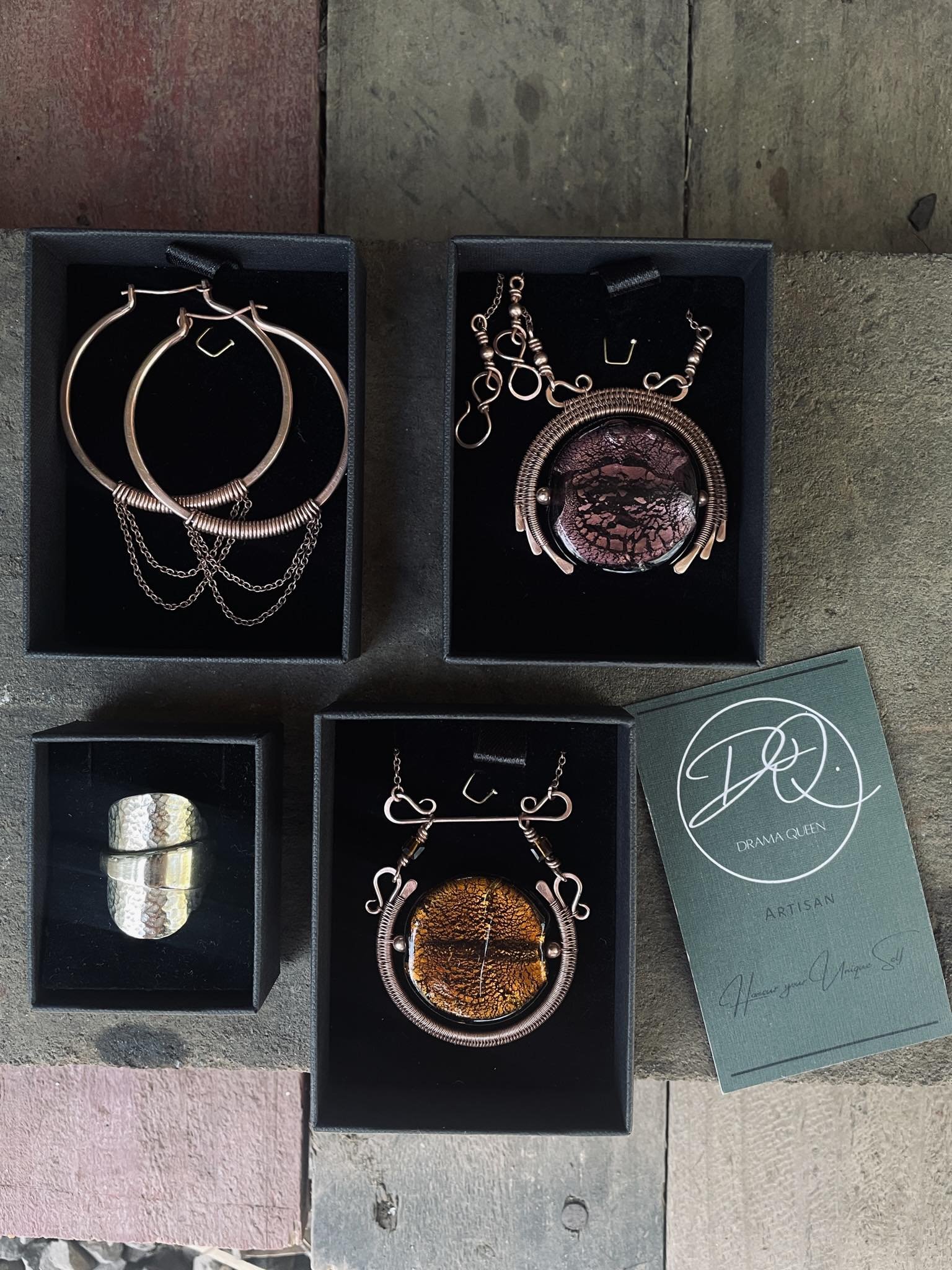 Four jewelry boxes containing necklaces, earrings, a ring, and a business card from Drama Queen Artisan, arranged on a concrete surface.