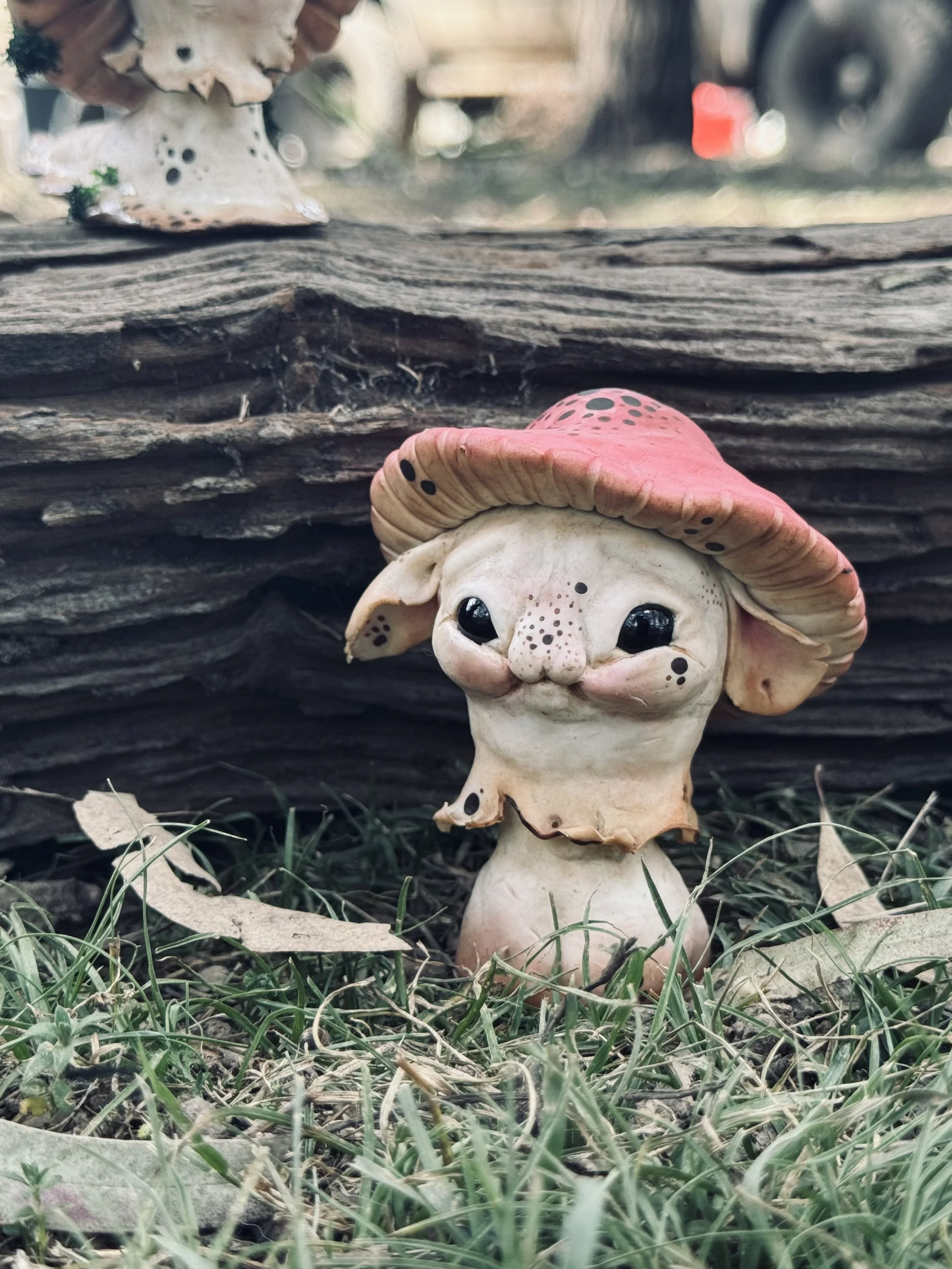 A whimsical mushroom sculpture with a cute, smiling face, black shiny eyes, and pink cap with black spots, set on grass in front of stacked wooden planks.
