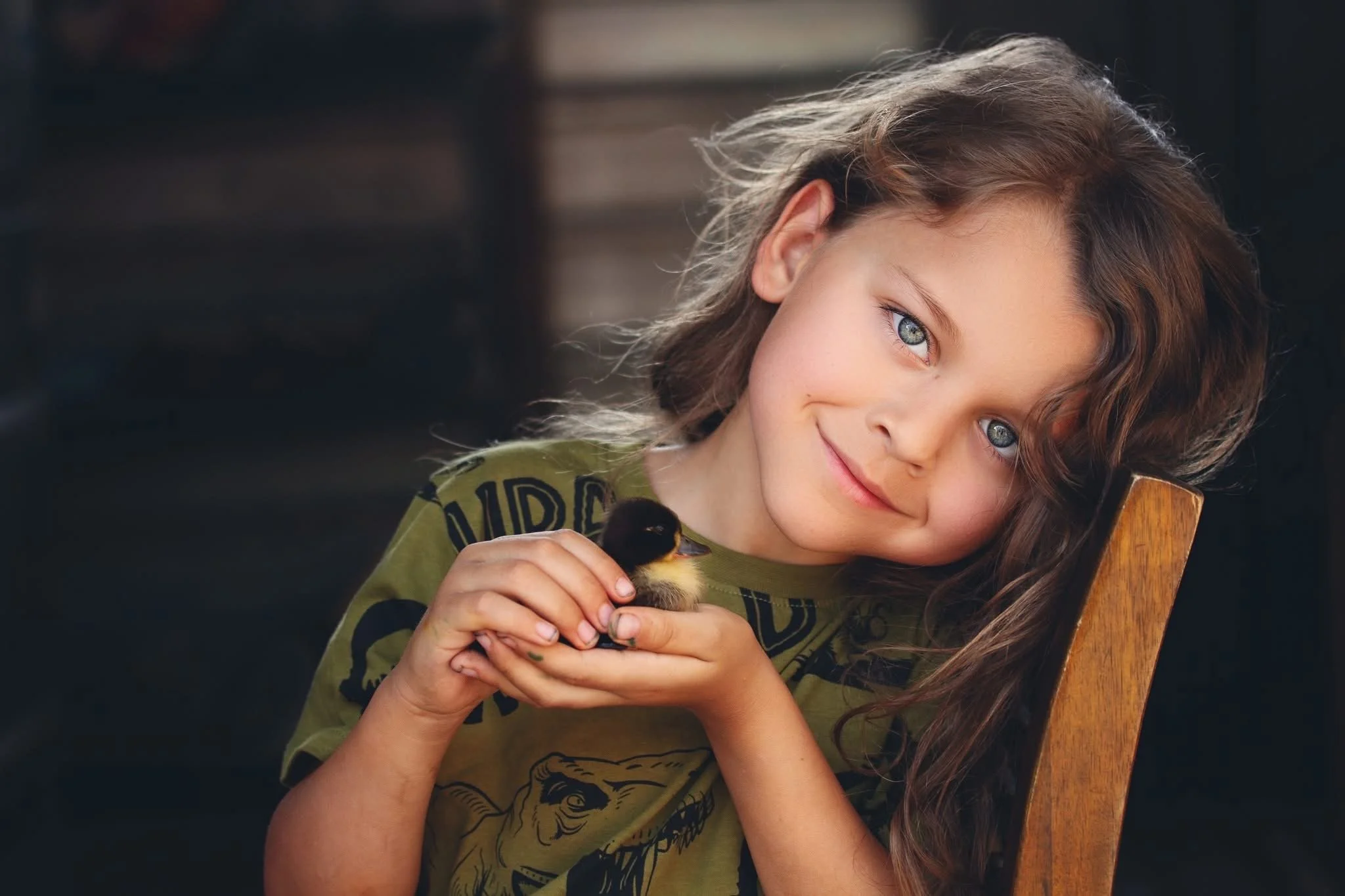 A young boy with long wavy brown hair and blue eyes holds a small duckling, smiling softly while resting hishead on a wooden chair back in a cozy indoor setting.