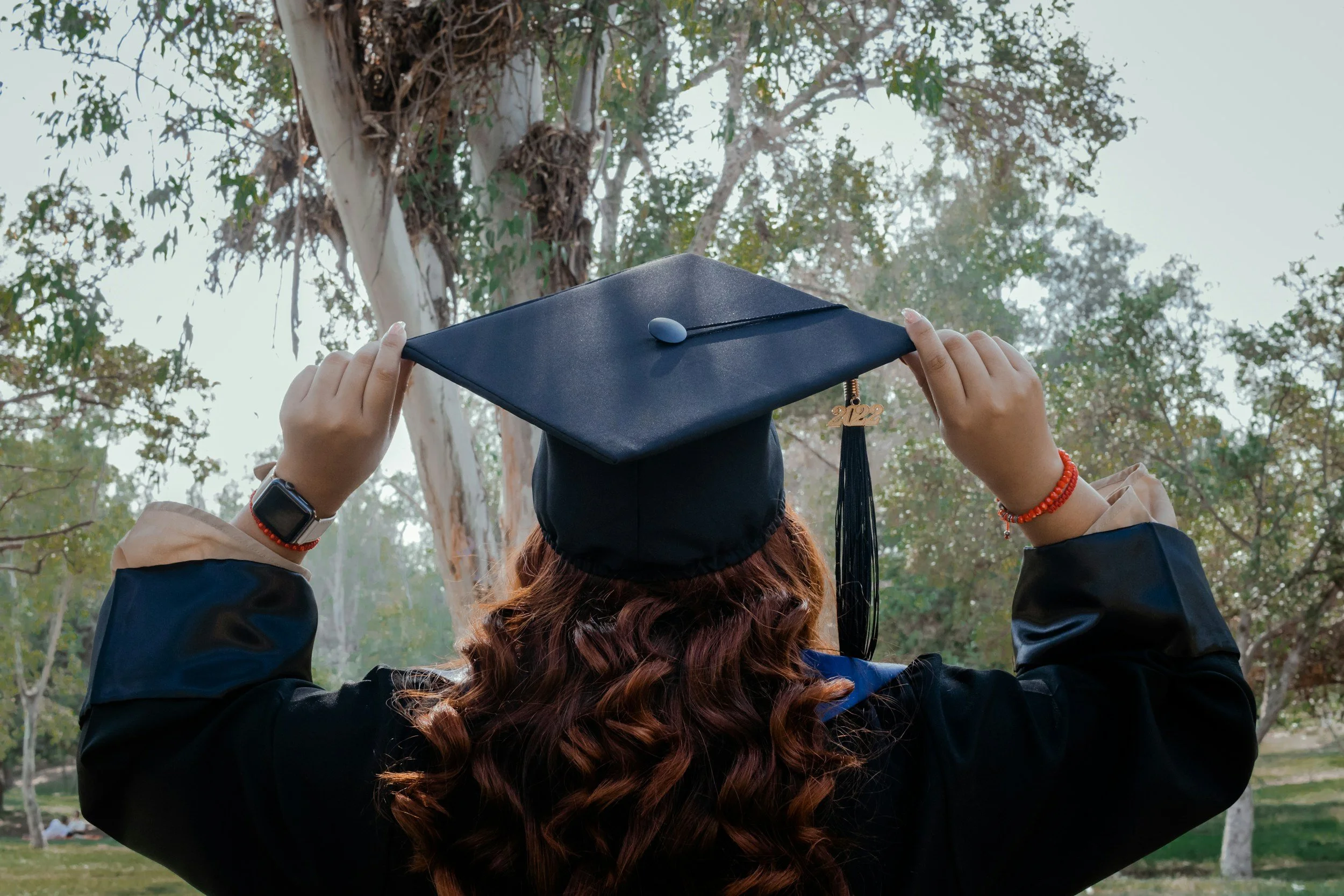 Person in graduation cap and gown adjusting the cap outdoors with trees in the background.