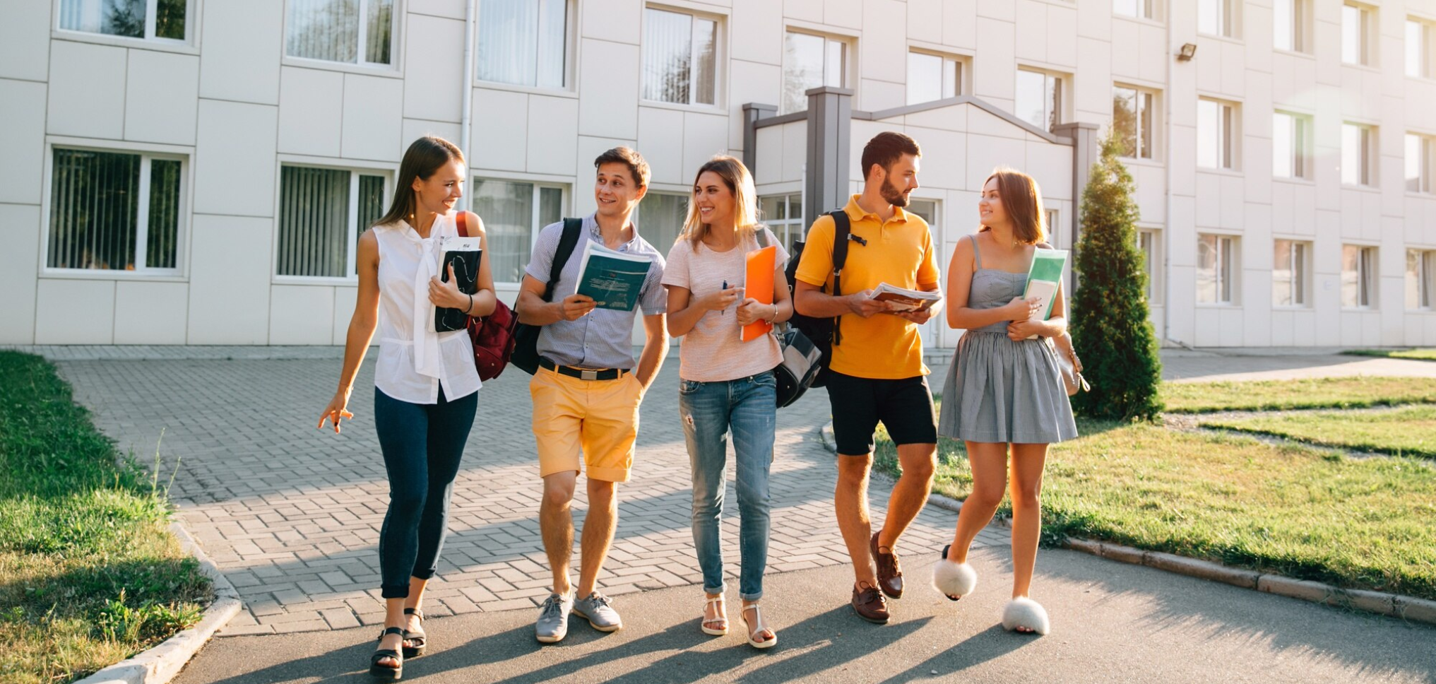 Group of five young students walking and talking outside a modern building, carrying books and backpacks on a sunny day.