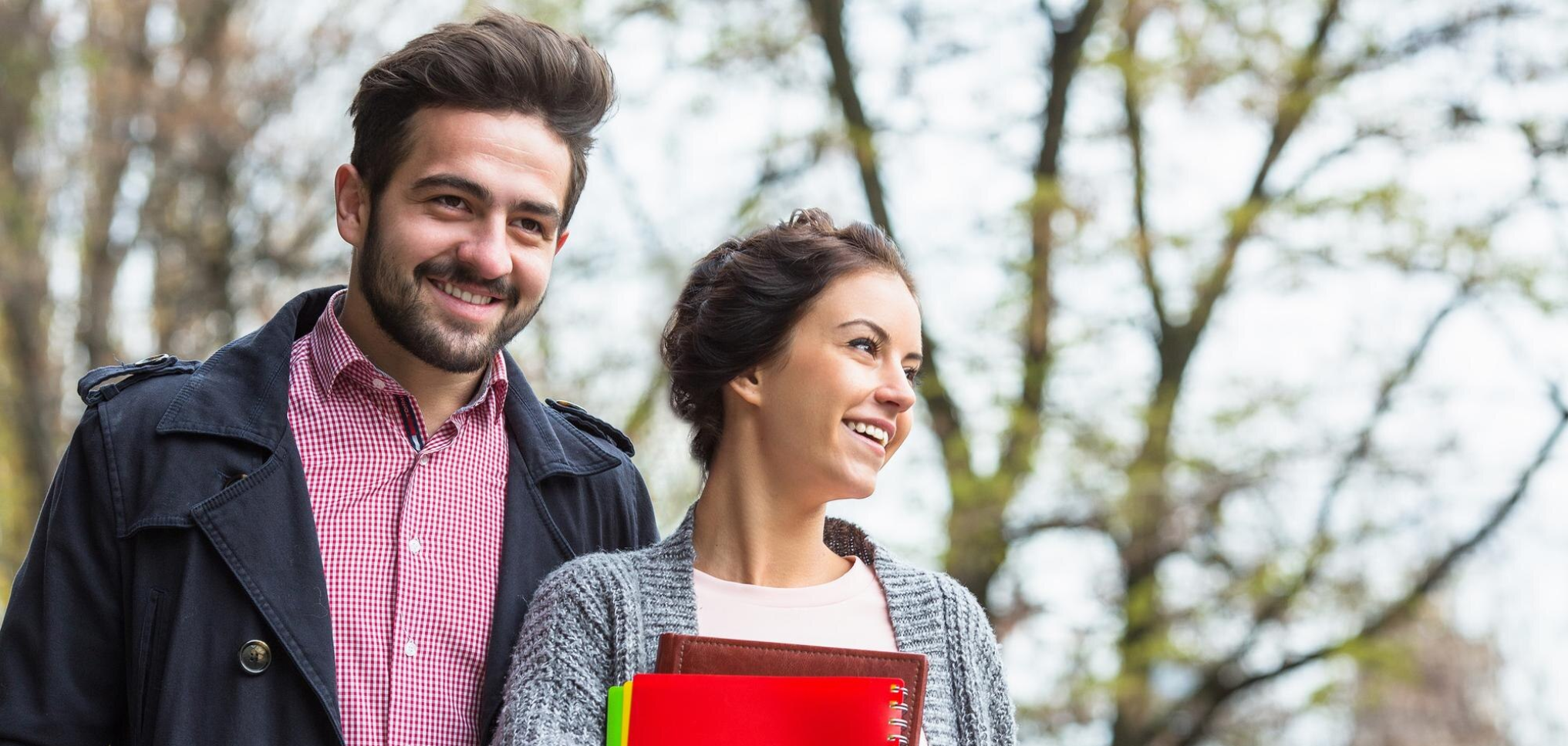 A young man and woman smiling outdoors in a park with trees in the background. The man has dark hair and a beard, wearing a dark jacket over a red and white checkered shirt. The woman has dark hair in a bun, wearing a gray knitted sweater, holding a red folder and notebook.