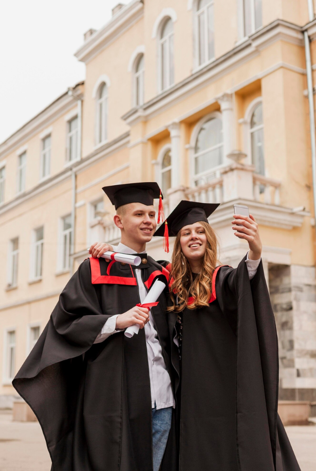 Two young graduates in black caps and gowns taking a selfie with a smartphone outside a large, historic building.