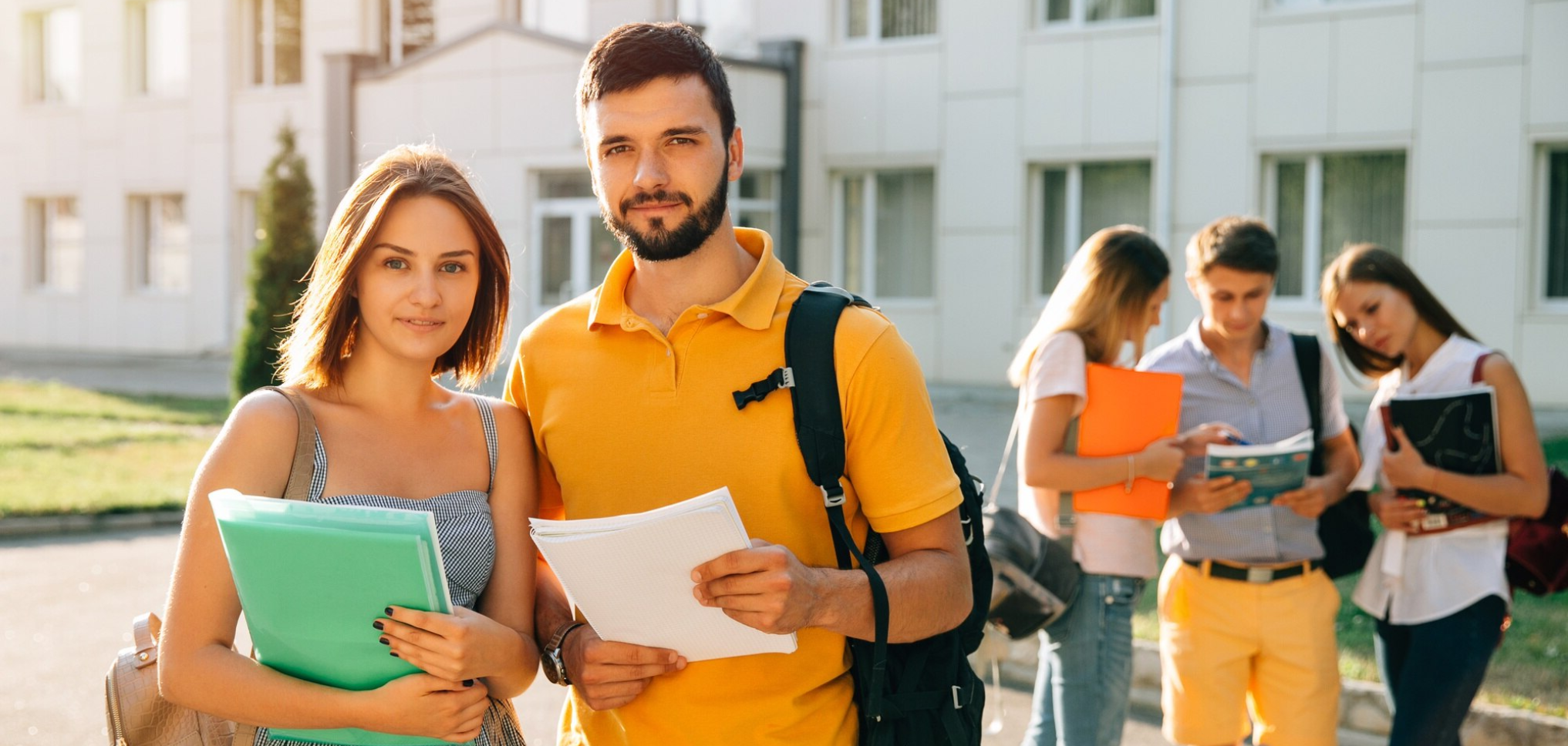 Group of young students standing outside school building during daytime, holding notebooks and folders, engaging with each other.