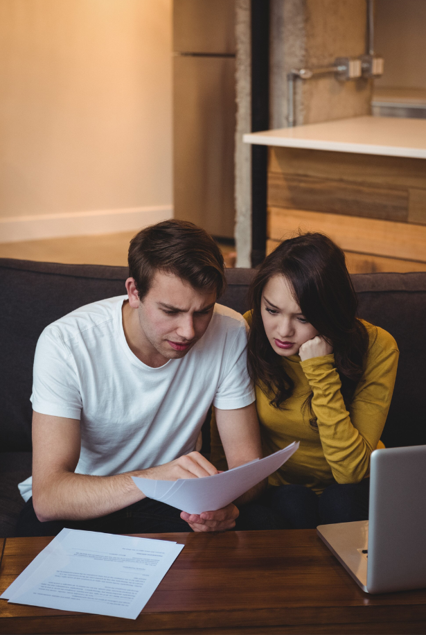 A young man and woman sitting on a couch looking at papers with a laptop and documents on the table, appearing worried.