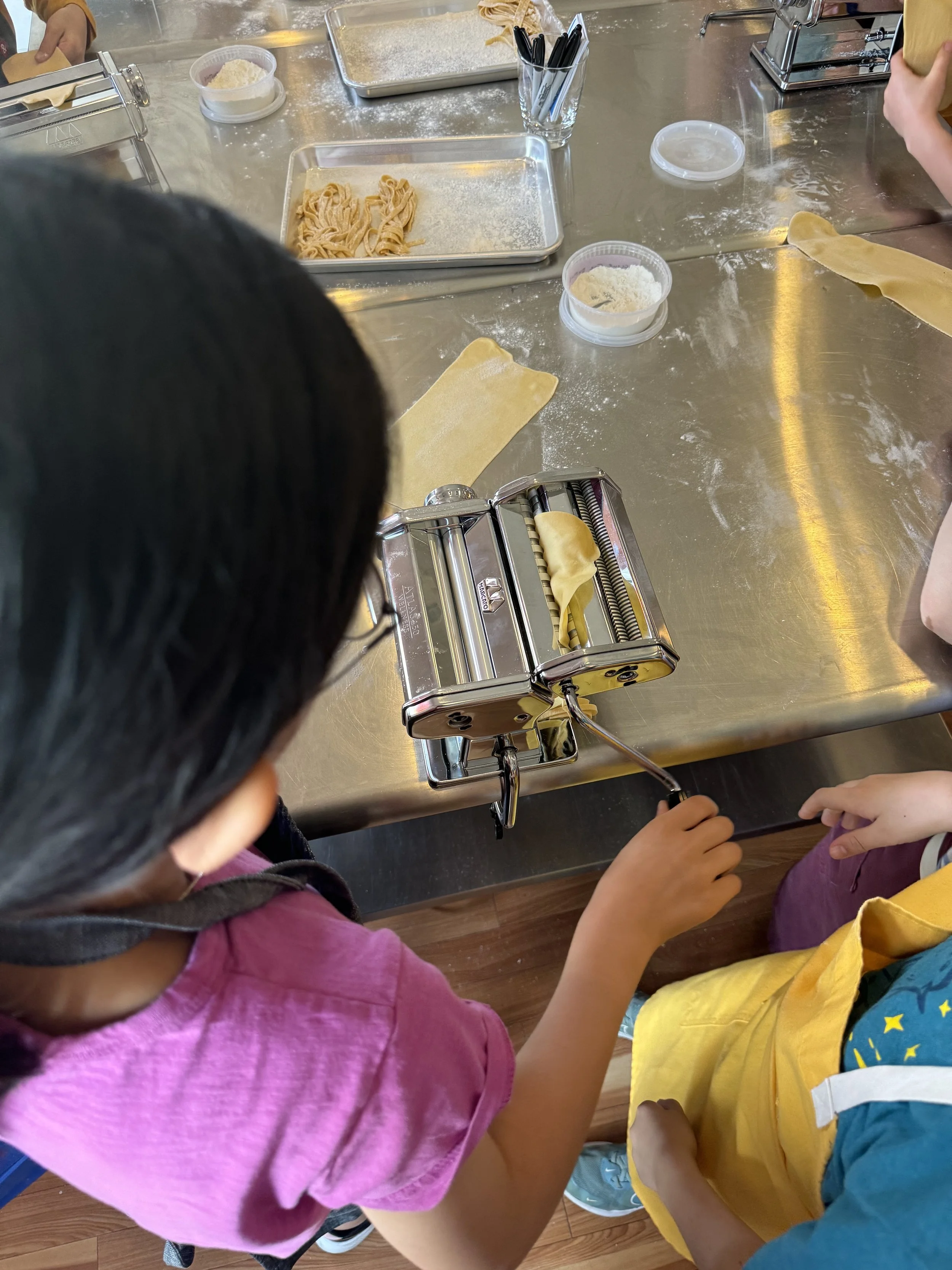 Children in a kitchen or baking class are making pasta, with a pasta machine, flour, and pasta dough on a metallic table.