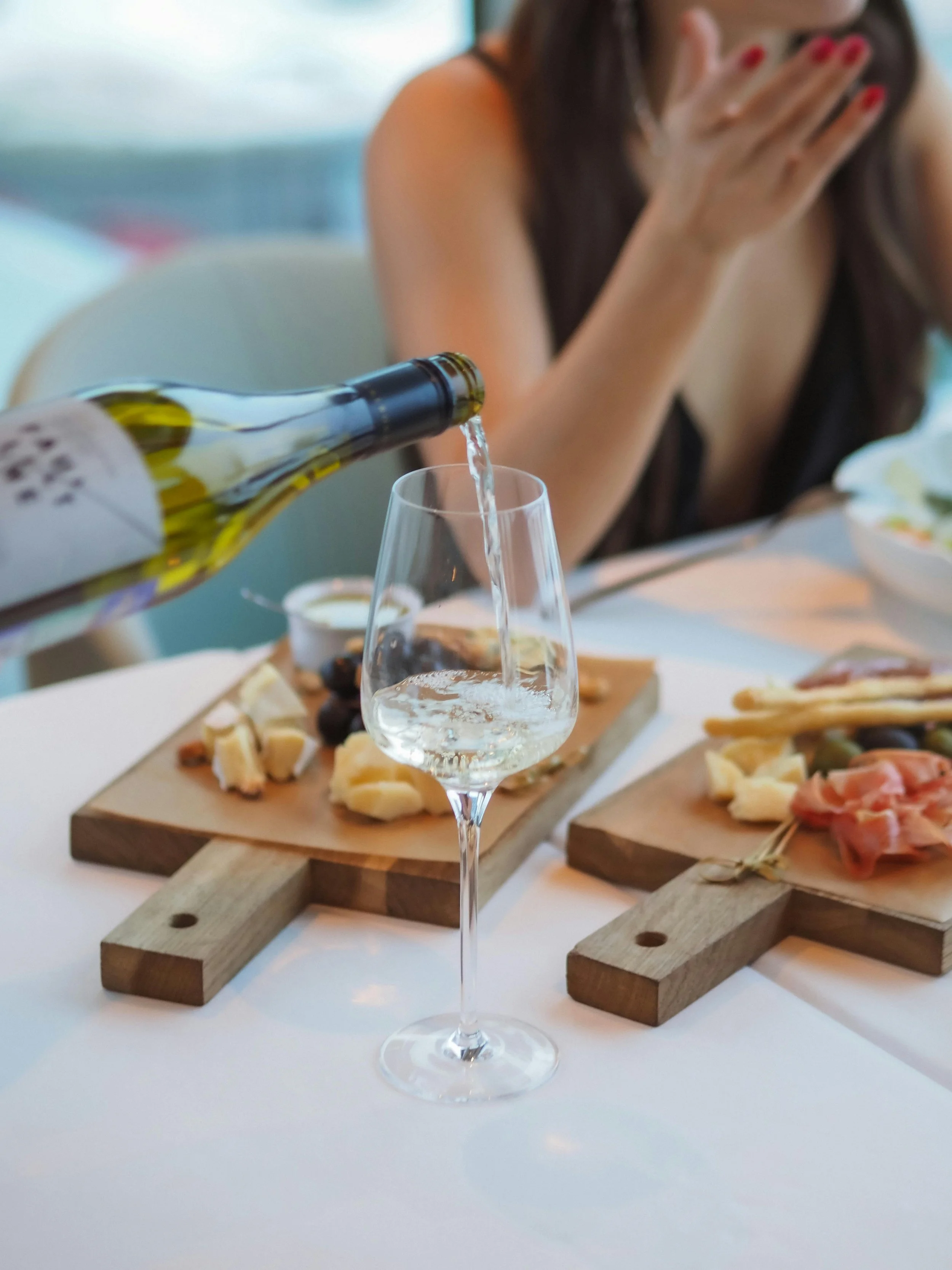 Person pouring white wine into a glass on a table set with cheese and charcuterie boards.