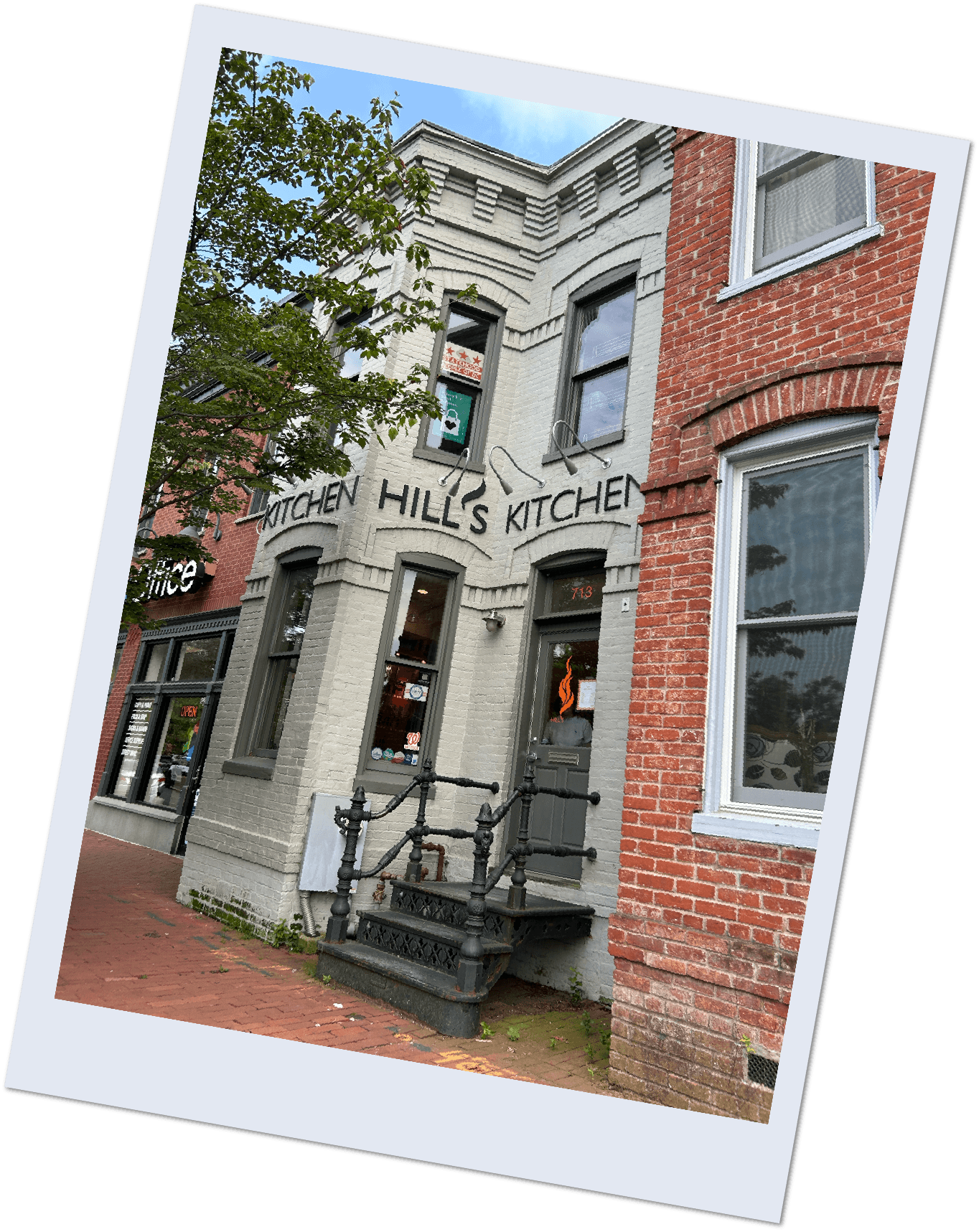 White brick building with black sign that reads 'Kitchen Hill's Kitchen' and black stairs leading to a door, next to a red brick building.