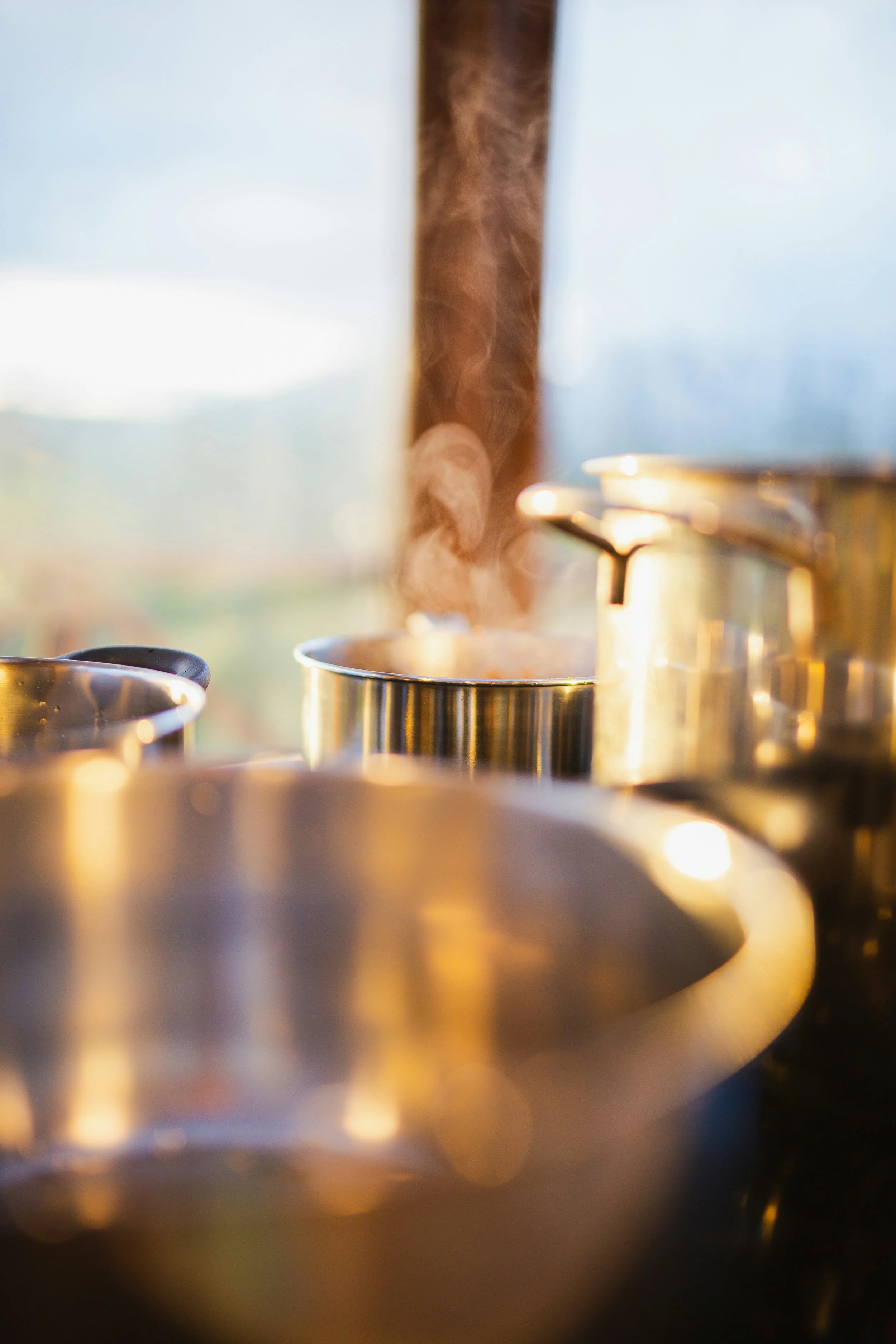 Close-up view of steaming pots on a stove, with sunlight shining through a window in the background.