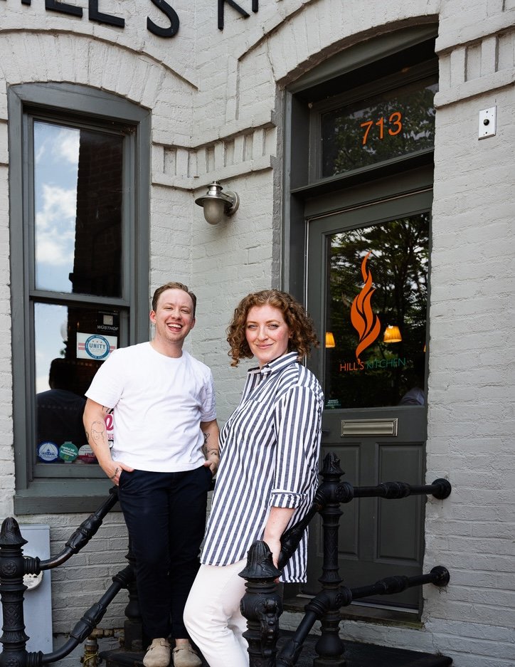 Two smiling women standing outside a restaurant with the number 713 on the door and a flame logo, one in a striped shirt and the other in a white T-shirt, in front of a gray brick building.