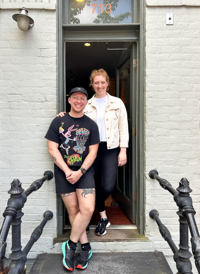 Two smiling women standing in a doorway of a white brick building. The woman on the left is wearing a black baseball cap, black T-shirt with cartoon characters, black shorts, and sneakers. The woman on the right is wearing a white jacket, white T-shirt, black pants, and sneakers.