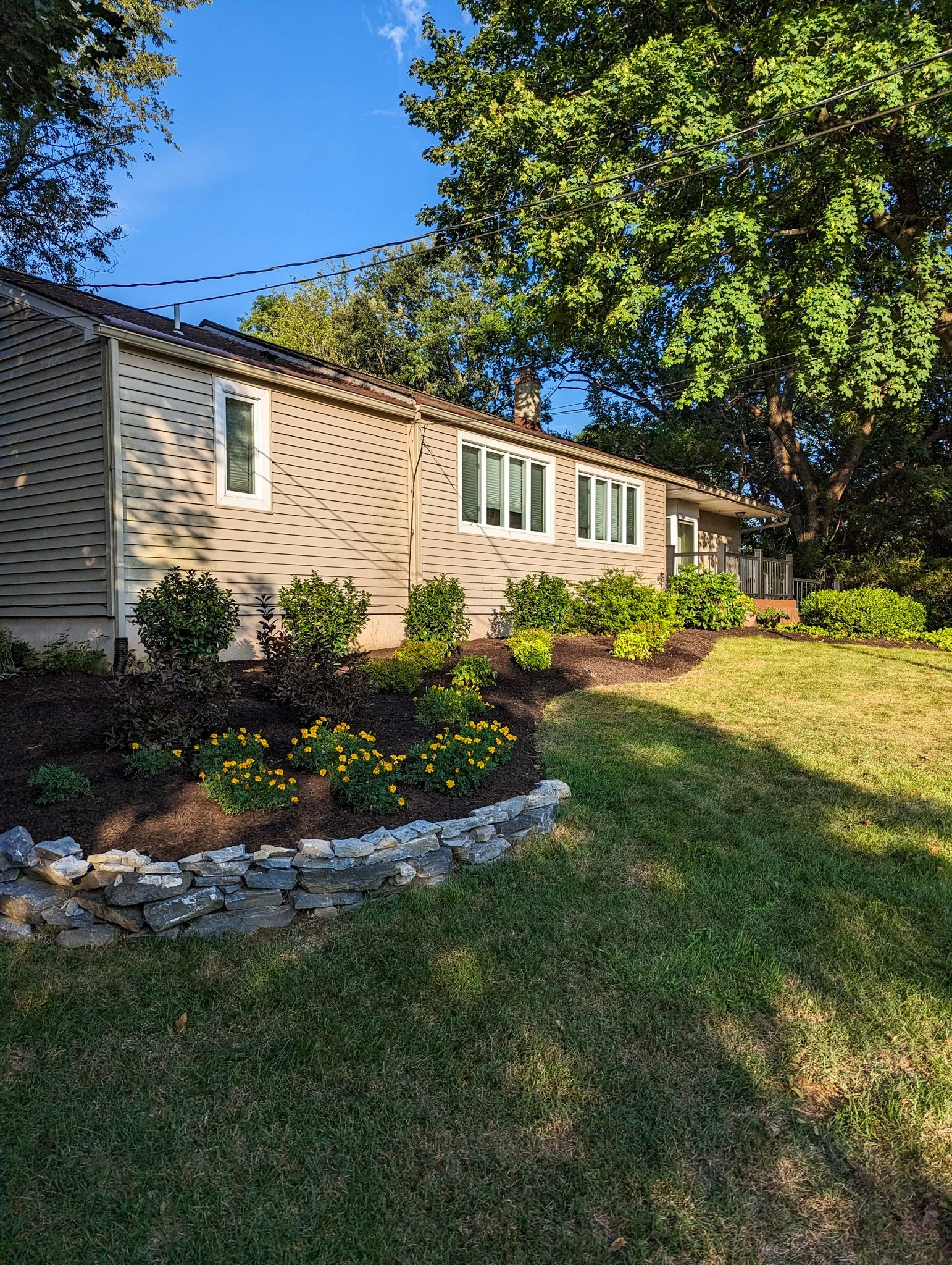 A beige house with white windows and a small porch, surrounded by green shrubbery and a freshly landscaped garden bed with yellow flowers, a stone border, and a well-maintained lawn, under a blue sky with some clouds.