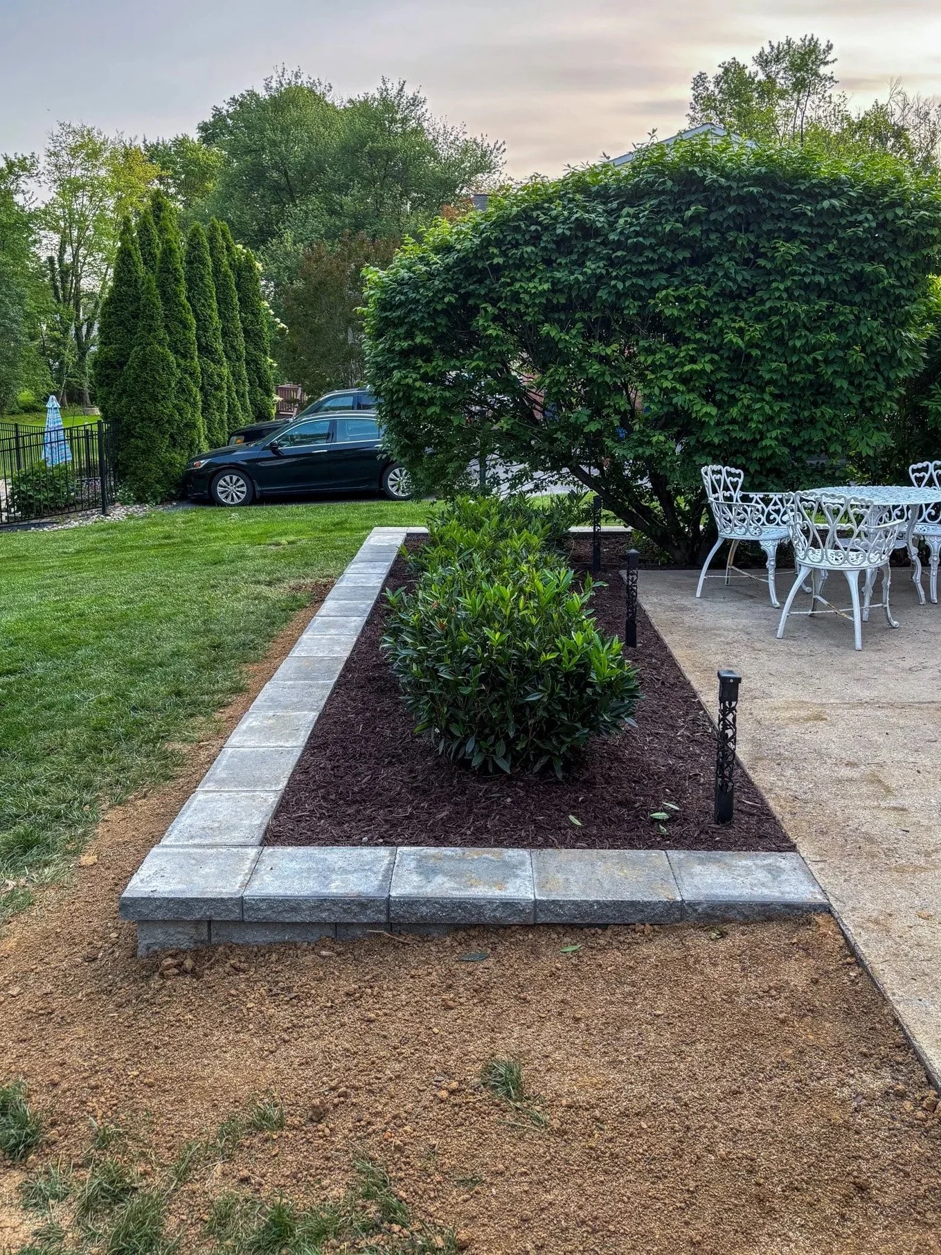 A landscaped backyard with a raised flower bed bordered by gray stone pavers, filled with dark mulch and green bushes. To the right, there is a concrete patio with white metal outdoor furniture and small pathway lights. In the background, a black car is parked near a row of tall, narrow evergreen trees, with a green lawn and large leafy trees further back.