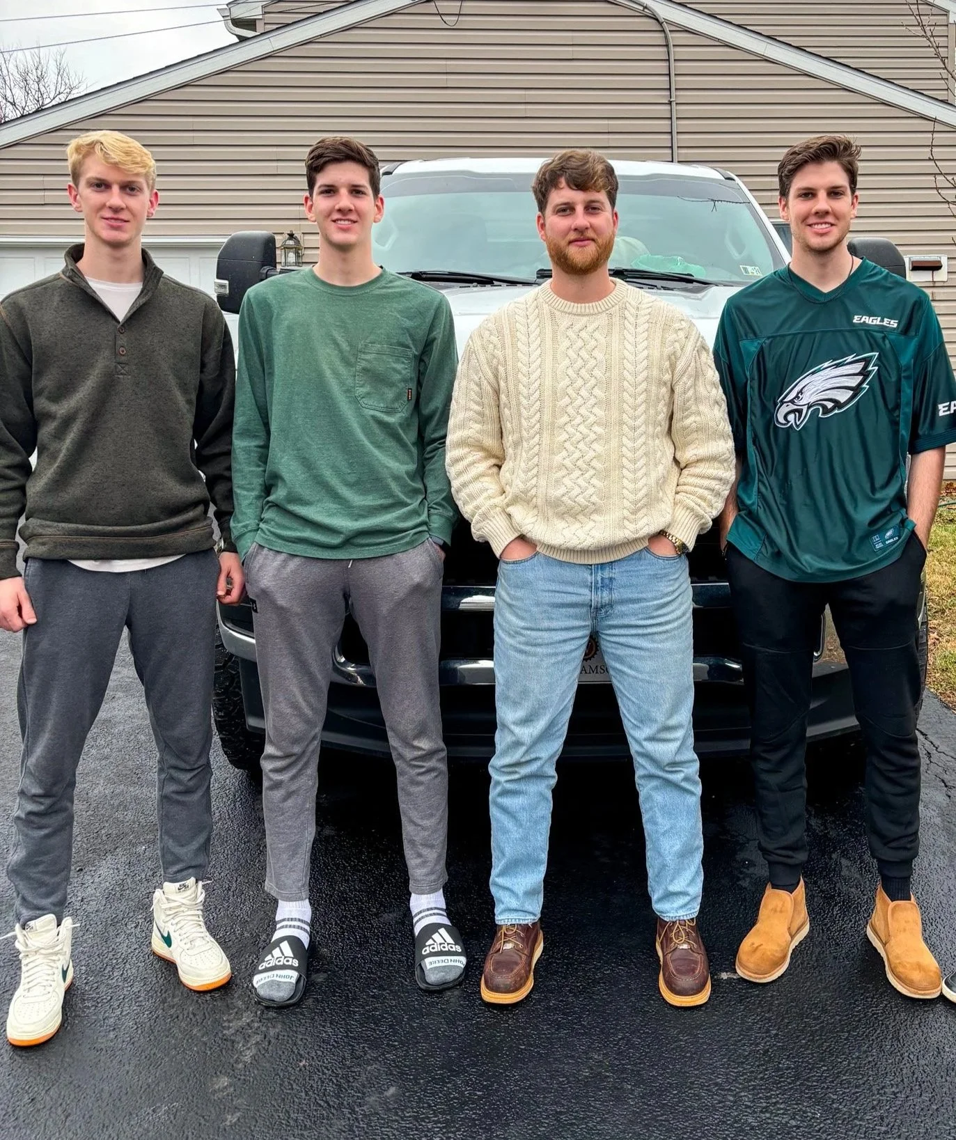 Four young men standing in front of a black vehicle, outdoors in a driveway, with a house in the background.