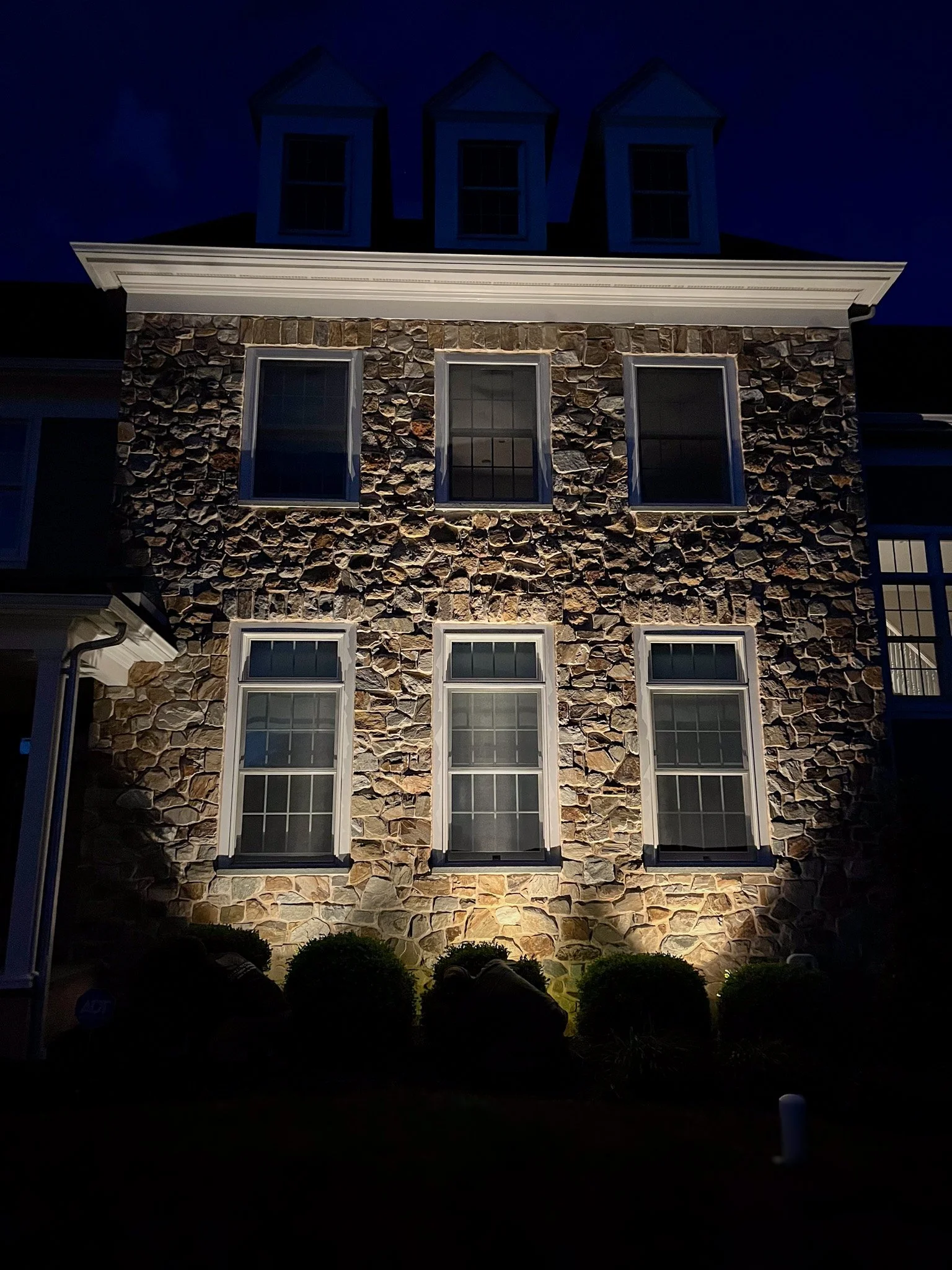 Front exterior of a multi-story house at night showing a stone facade, six windows with white trim, and bushes at the base illuminated by outdoor lighting.