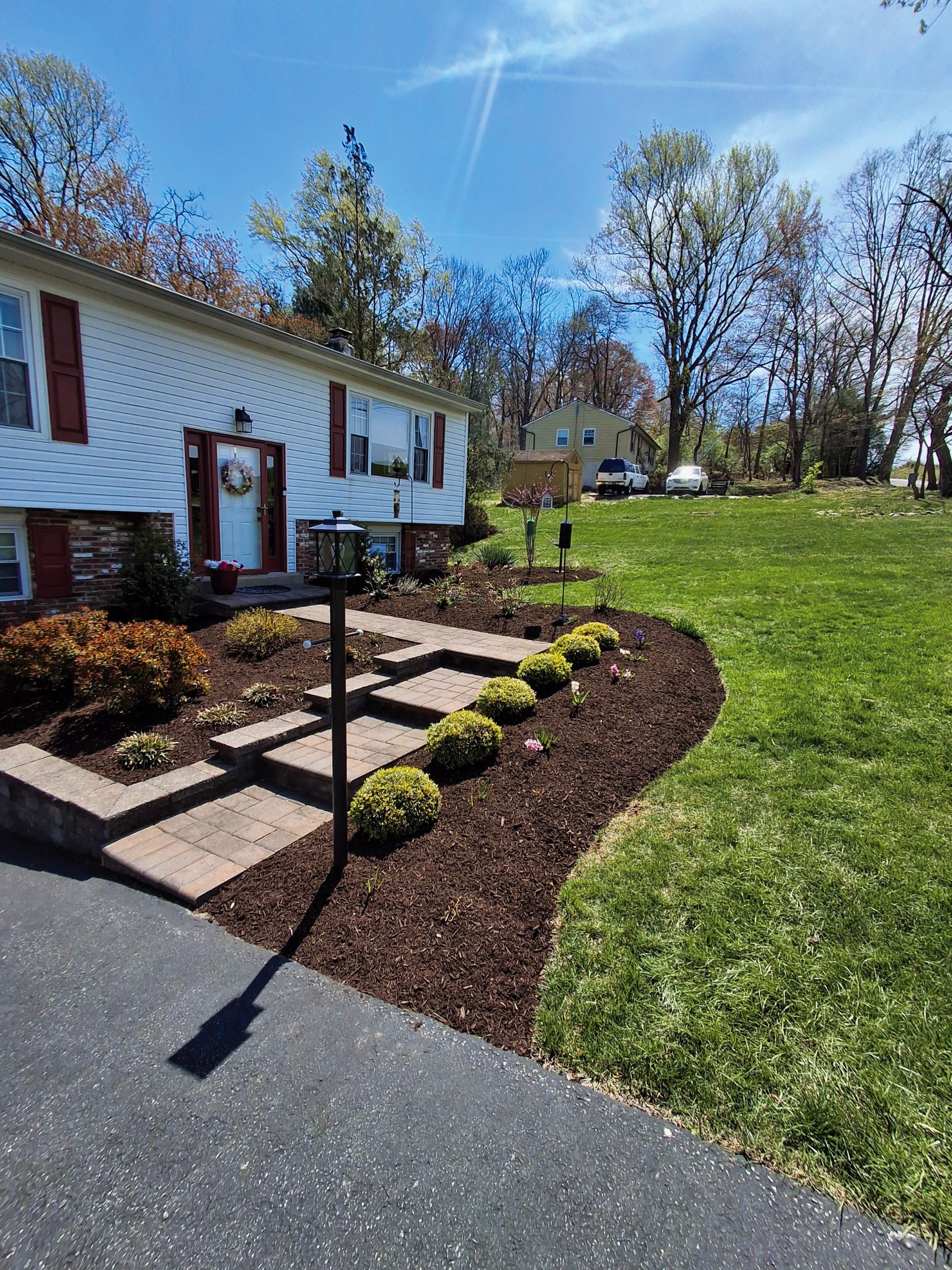 Front yard with a landscaped garden, stone pathway, lamp posts, a white house with red shutters and door wreath, green lawn, trees, and parked cars in the background.