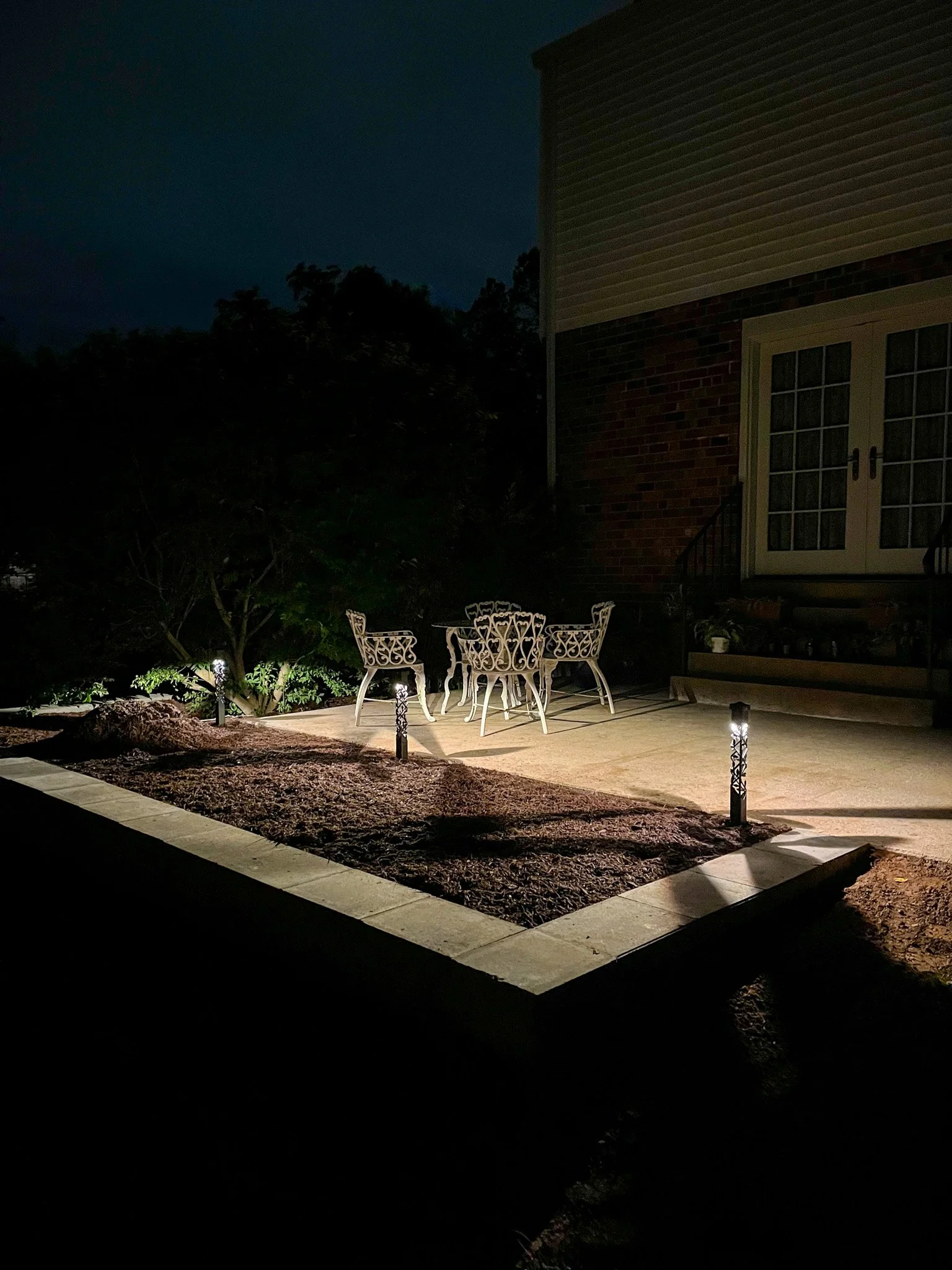Nighttime backyard patio with white ornate metal table and chairs, illuminated by landscape lighting, with a house featuring a sliding glass door and brick exterior wall in the background.