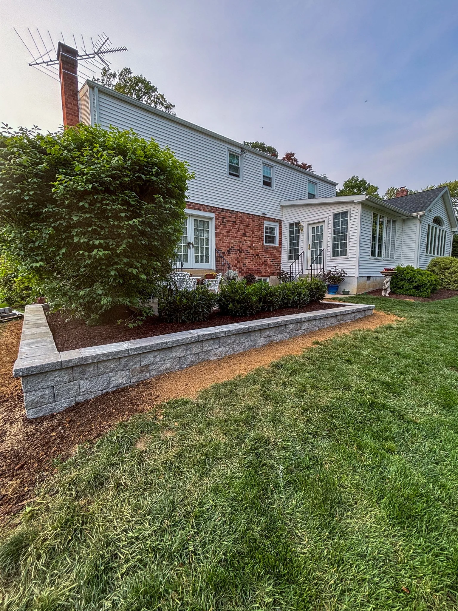 View of a backyard garden with a raised stone flower bed, green shrubs, a tree, and a two-story house with white siding and brick accents.