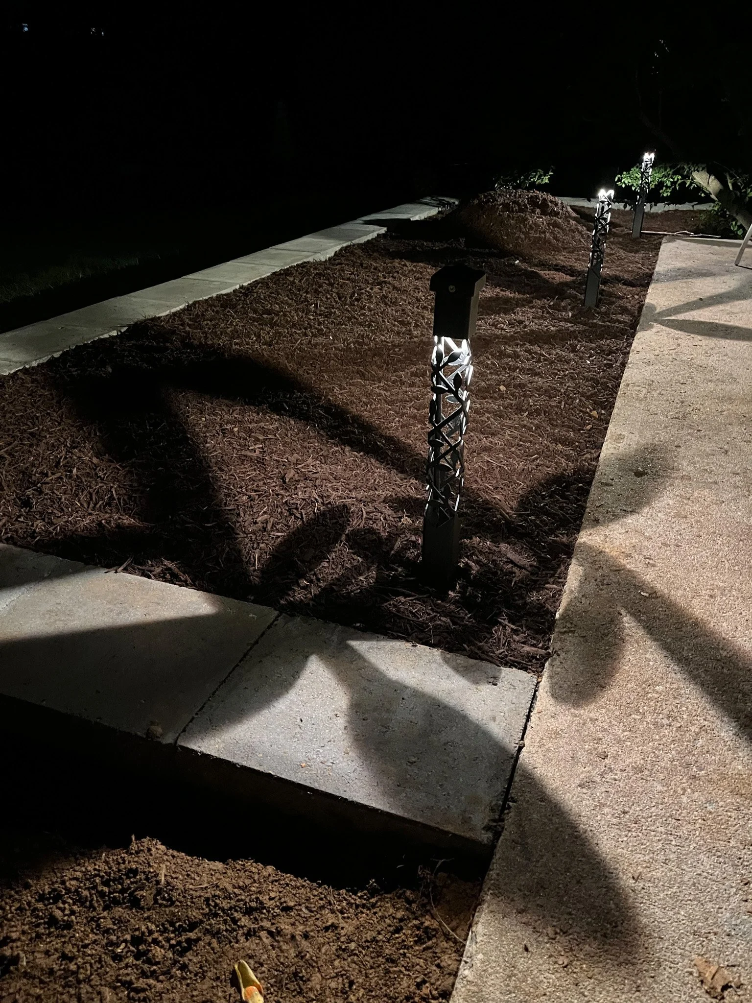 Nighttime outdoor scene featuring a landscaped area with a mulch bed, decorative lighting fixtures, and a concrete sidewalk.